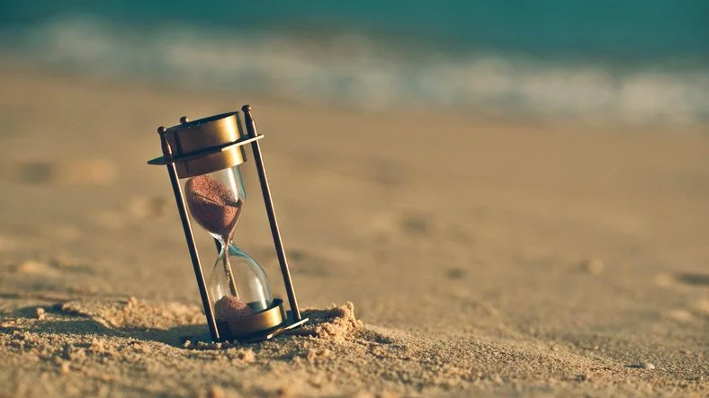 An old-fashioned hourglass nestled in the sand at the beach, with the water out of focus in the background