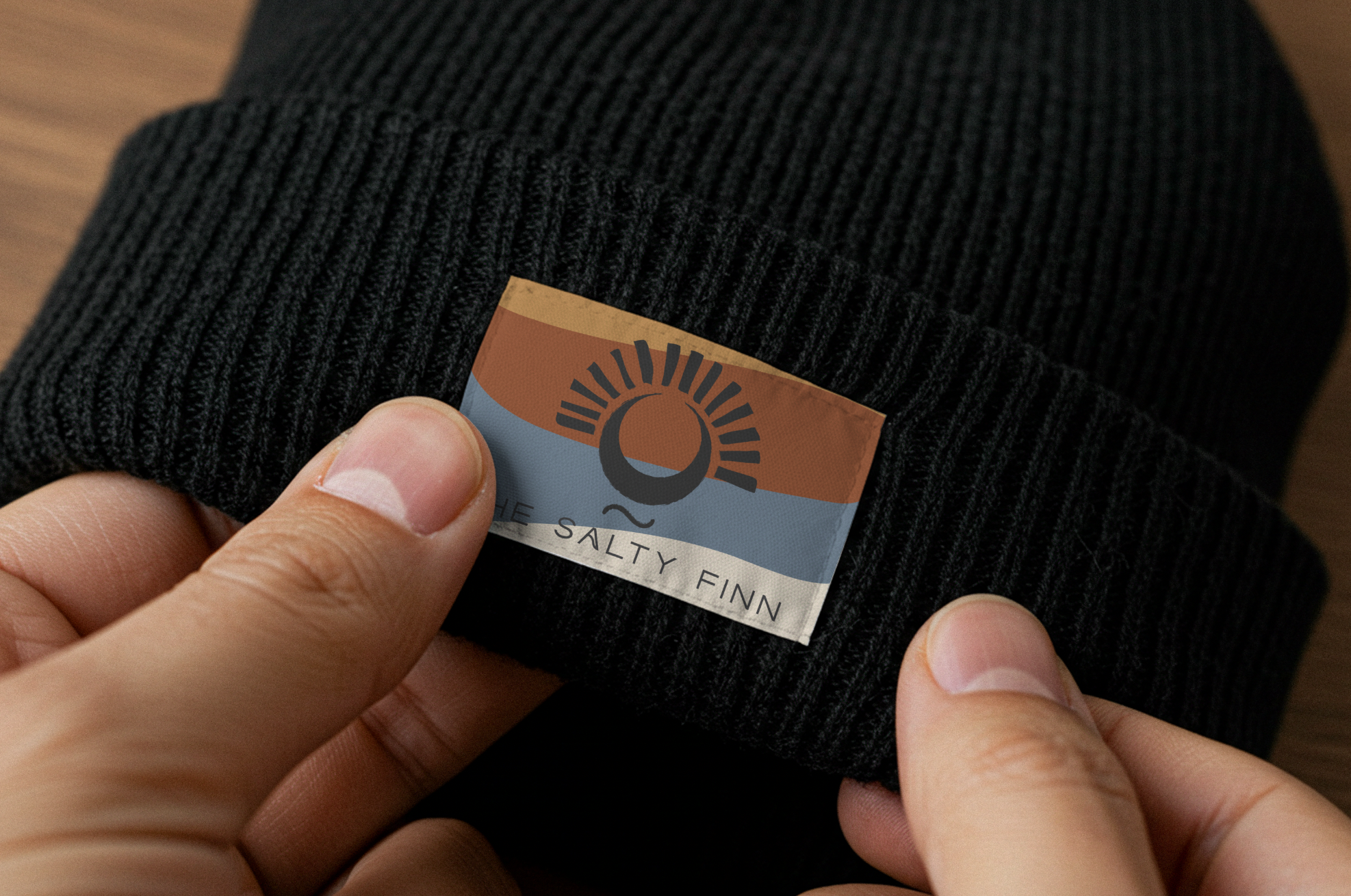 Close-up of a person's hand pulling back a black knit hat to reveal a tag that says 'The Salty Finn' with a logo of a sun over water.