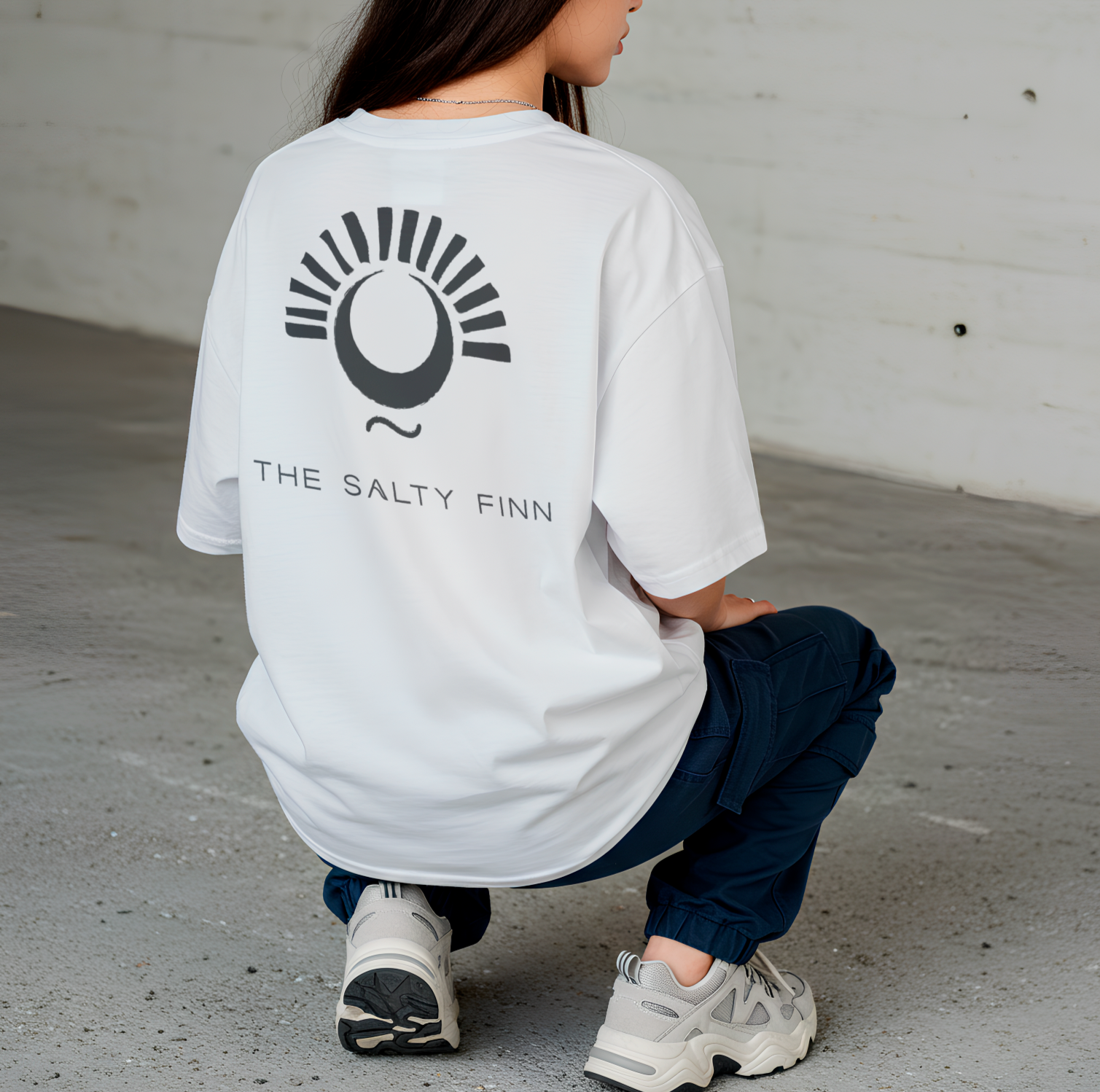 A person squatting wearing a white t-shirt with a graphic and the text "The Salty Finn," navy pants, and beige sneakers, against a white brick wall background.
