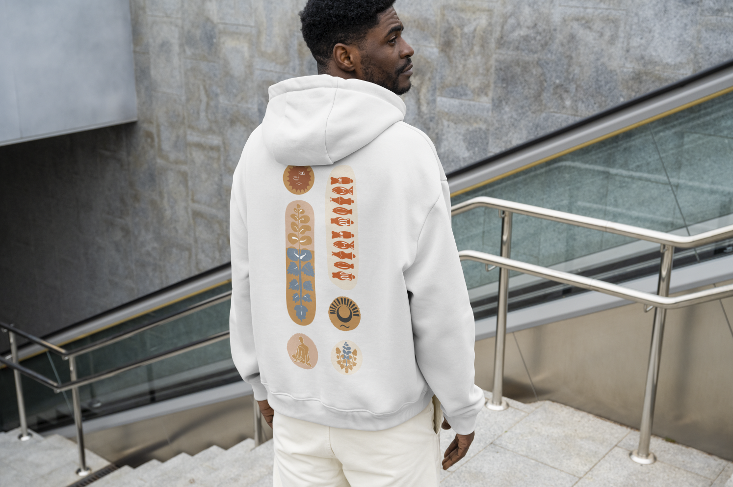 A man standing on stairs near a glass railing, wearing a white hoodie with decorative designs on the back.