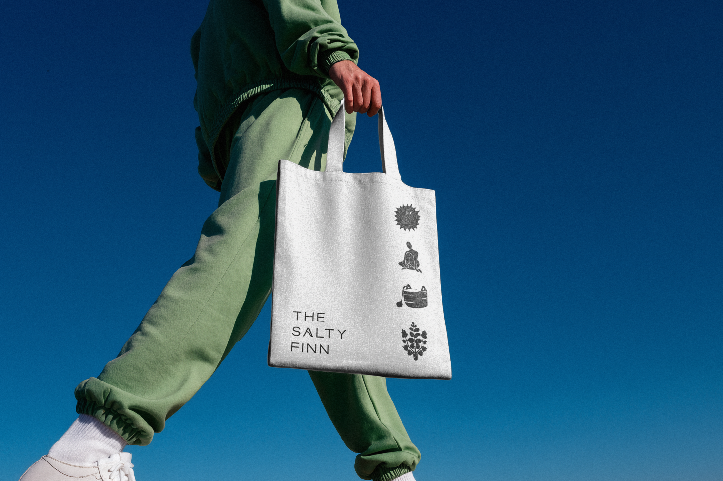 Person wearing light green pants and a green jacket holding a white tote bag with black icons and text, against a clear blue sky.
