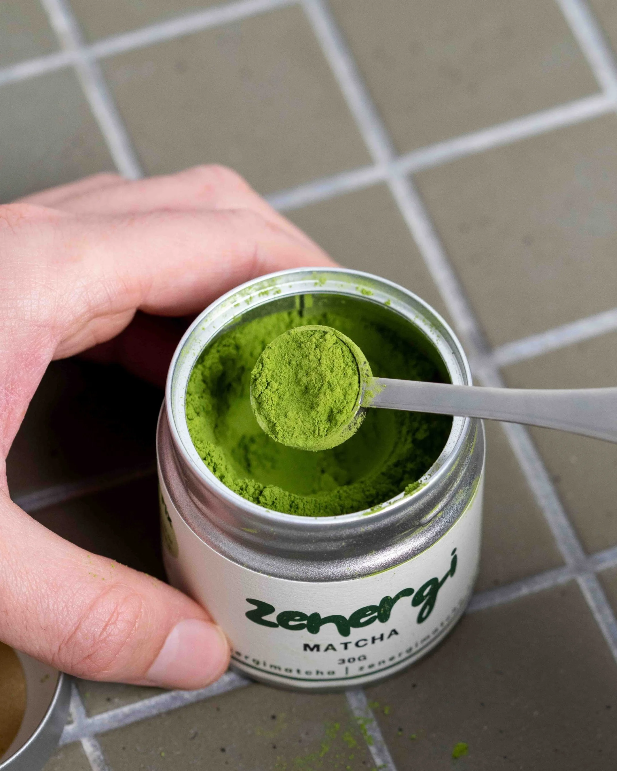 Open container of green matcha powder labeled 'Zenergy Matcha 30g' with a white scoop resting inside, showing bright green powdered matcha on a tiled surface.