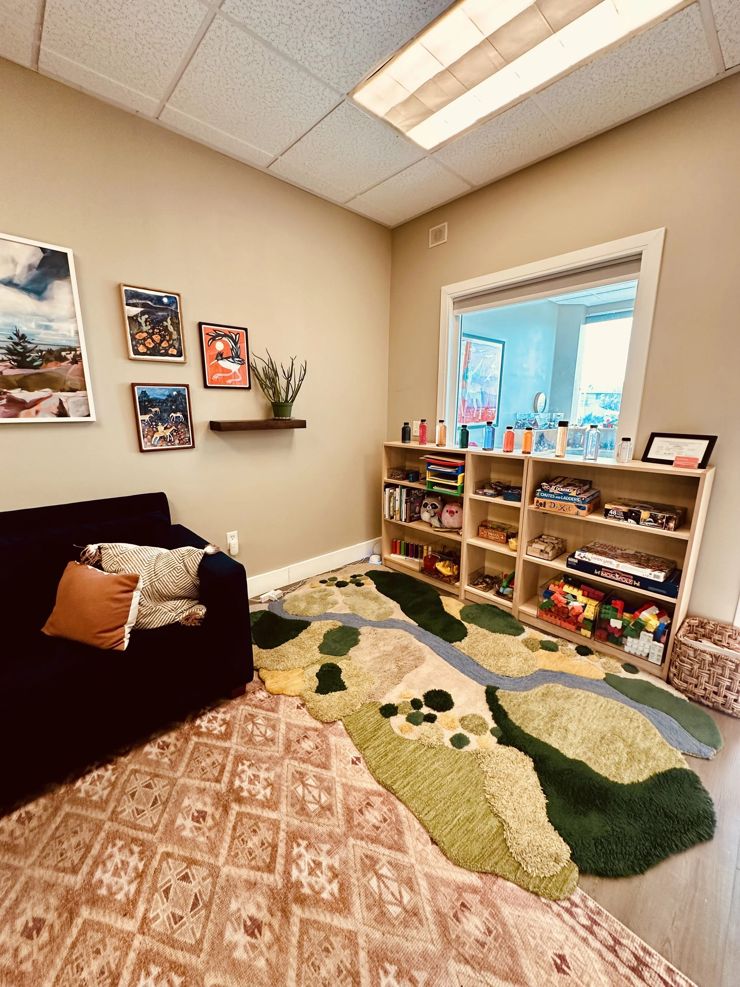 Lobby showing the end of a blue couch with desert themed pictures above. Bookshelves with toys and books along the next wall. The floor has a warm colored rug under the couch and a green and blue play rug by the bookshelves.