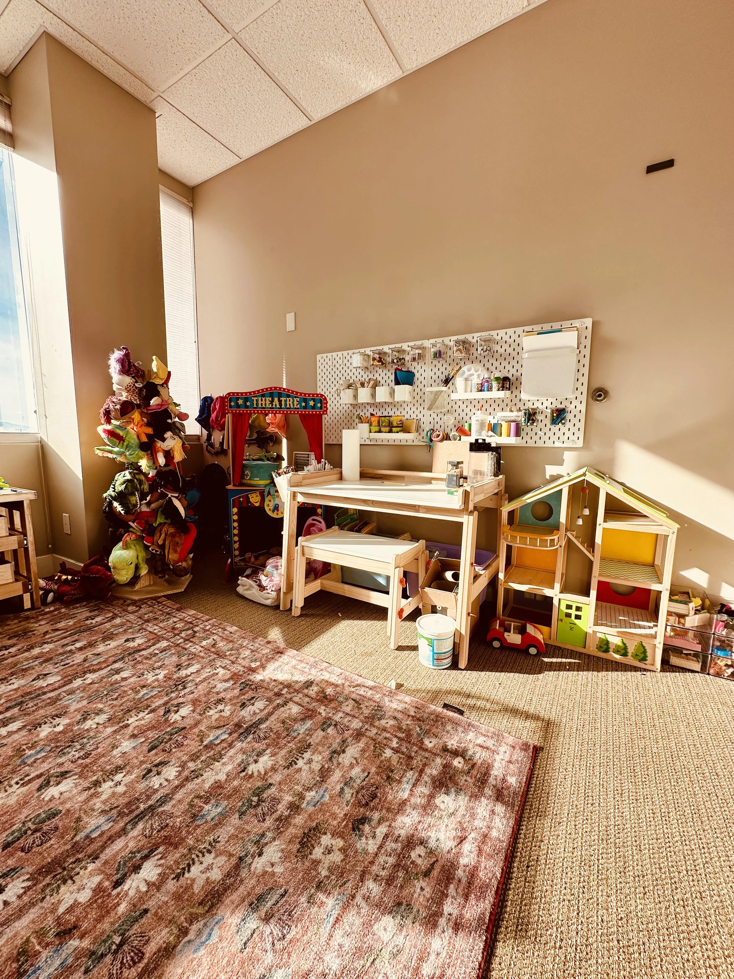 puppet stand with many colorful puppets on it. play theater beside this with an art table and art supplies on the wall above the table. play dollhouse on the ground next to the art station.