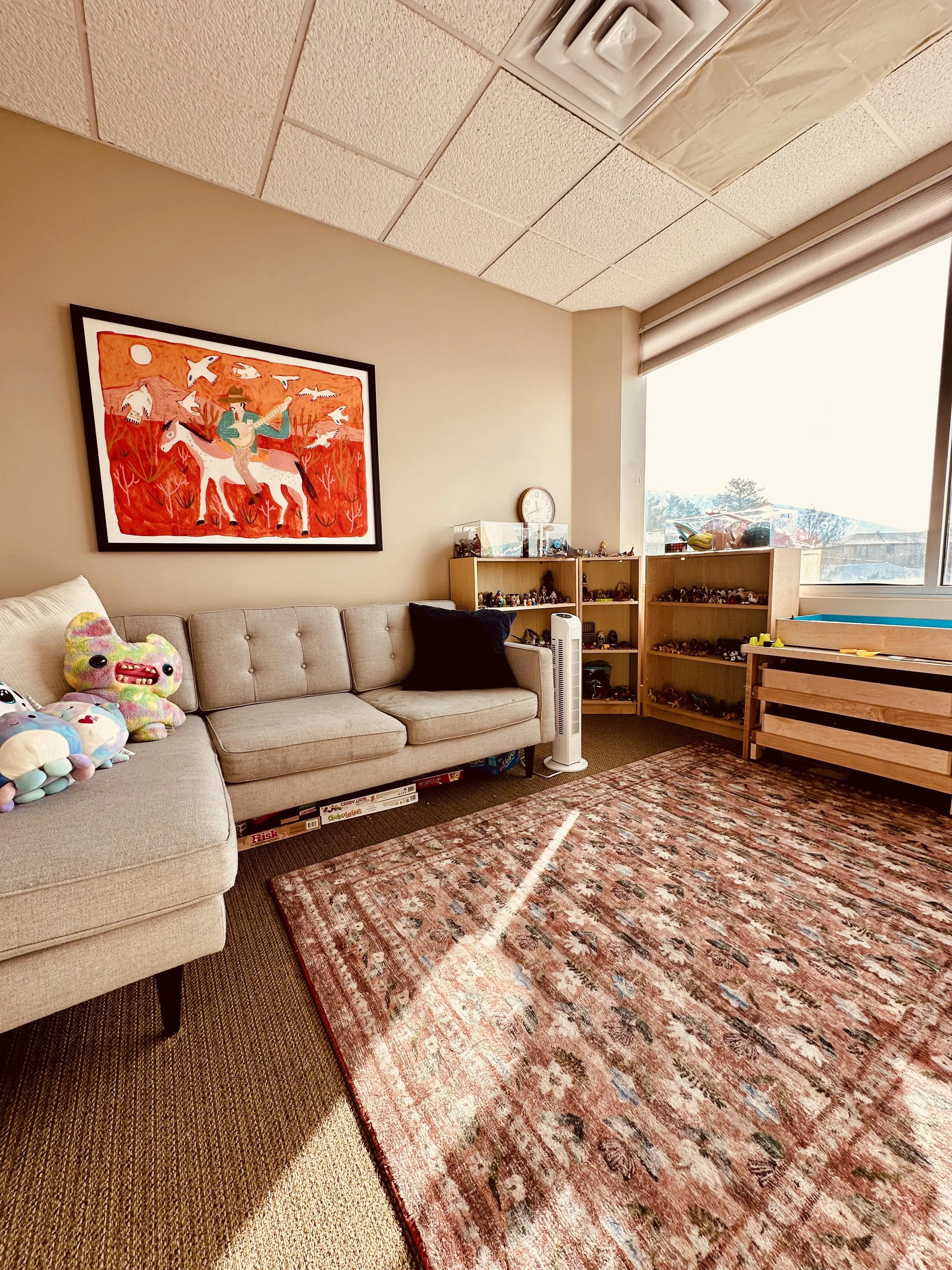 a grey couch with multiple pillows on it. a red and orange picture above the couch with a person riding a horse. bookshelves in the corner with toys and figures on them. warm colored rug