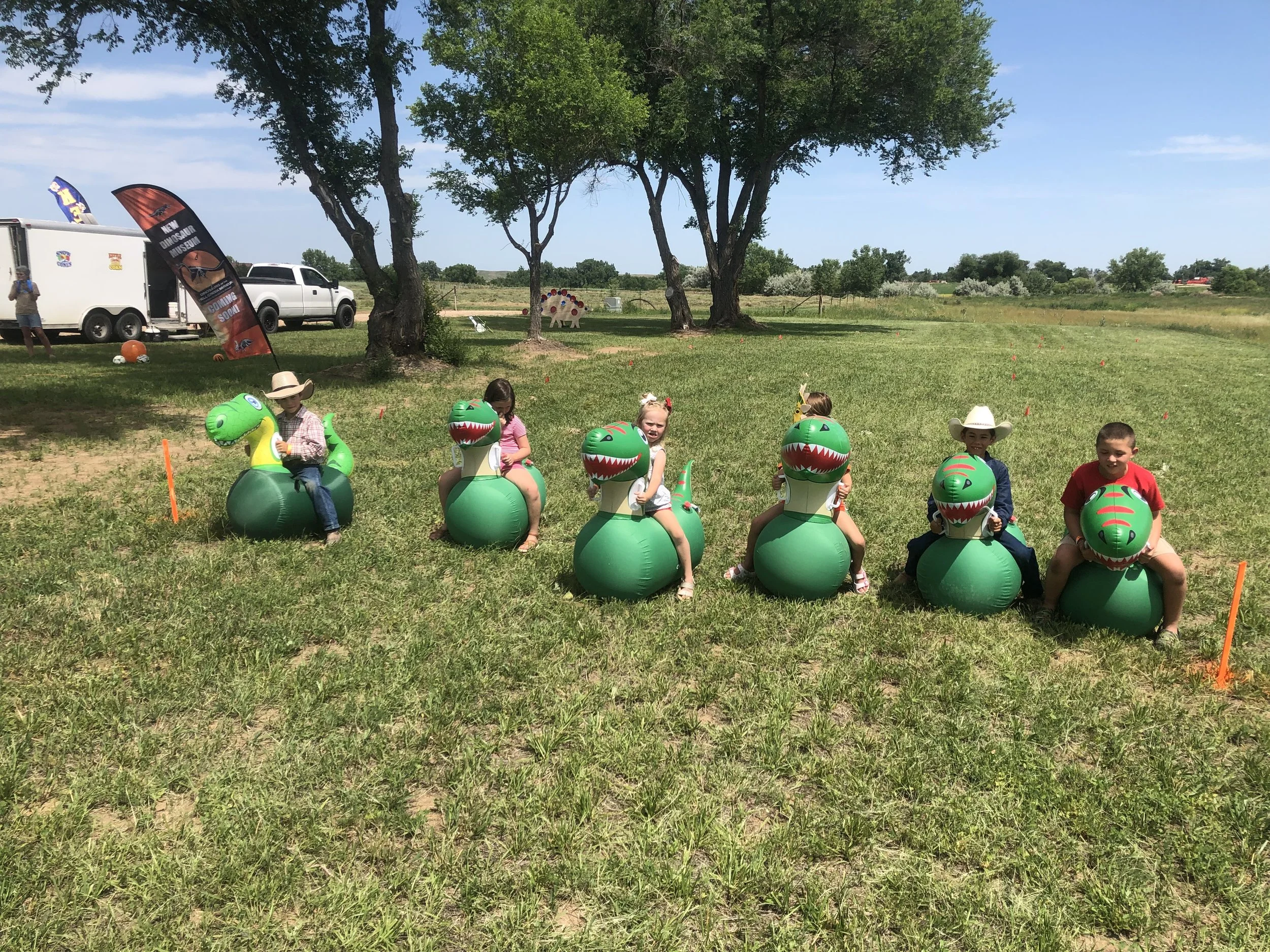 Person riding a large dinosaur fossil replica with an open mouth, wearing a wide-brimmed hat, blue shirt, and jeans, in an outdoor setting