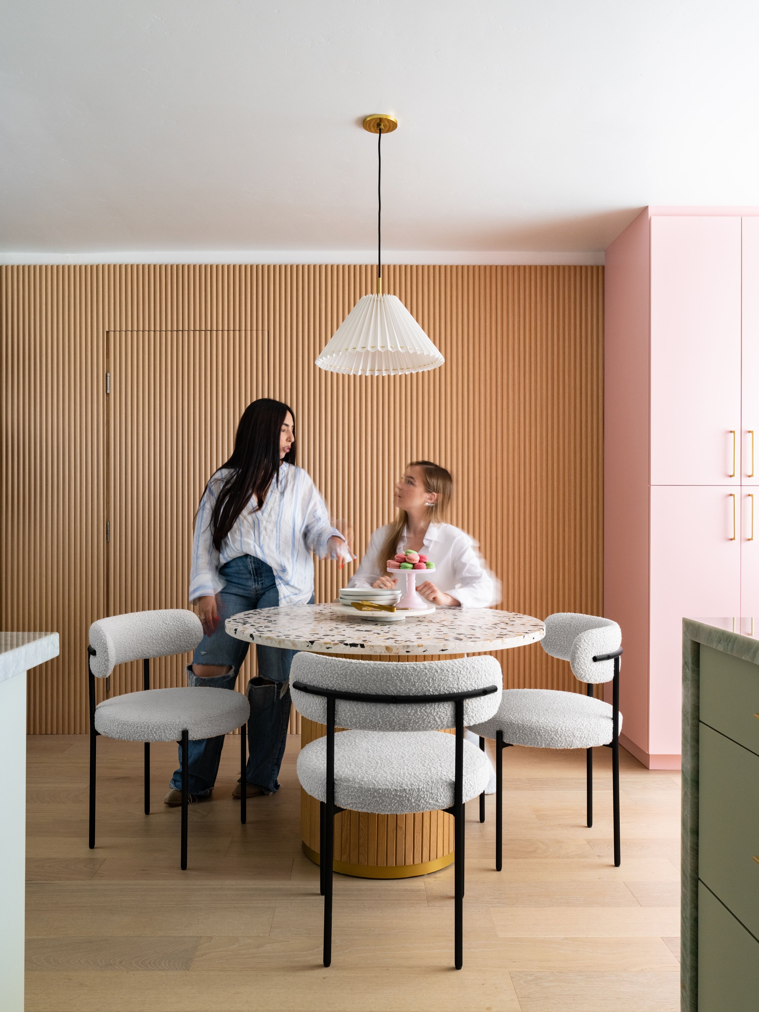 Two people in a modern kitchen with a marble table and stylish chairs. The room has light wooden flooring, pink cabinets, and a wooden slat wall. A pendant lamp hangs above the table.