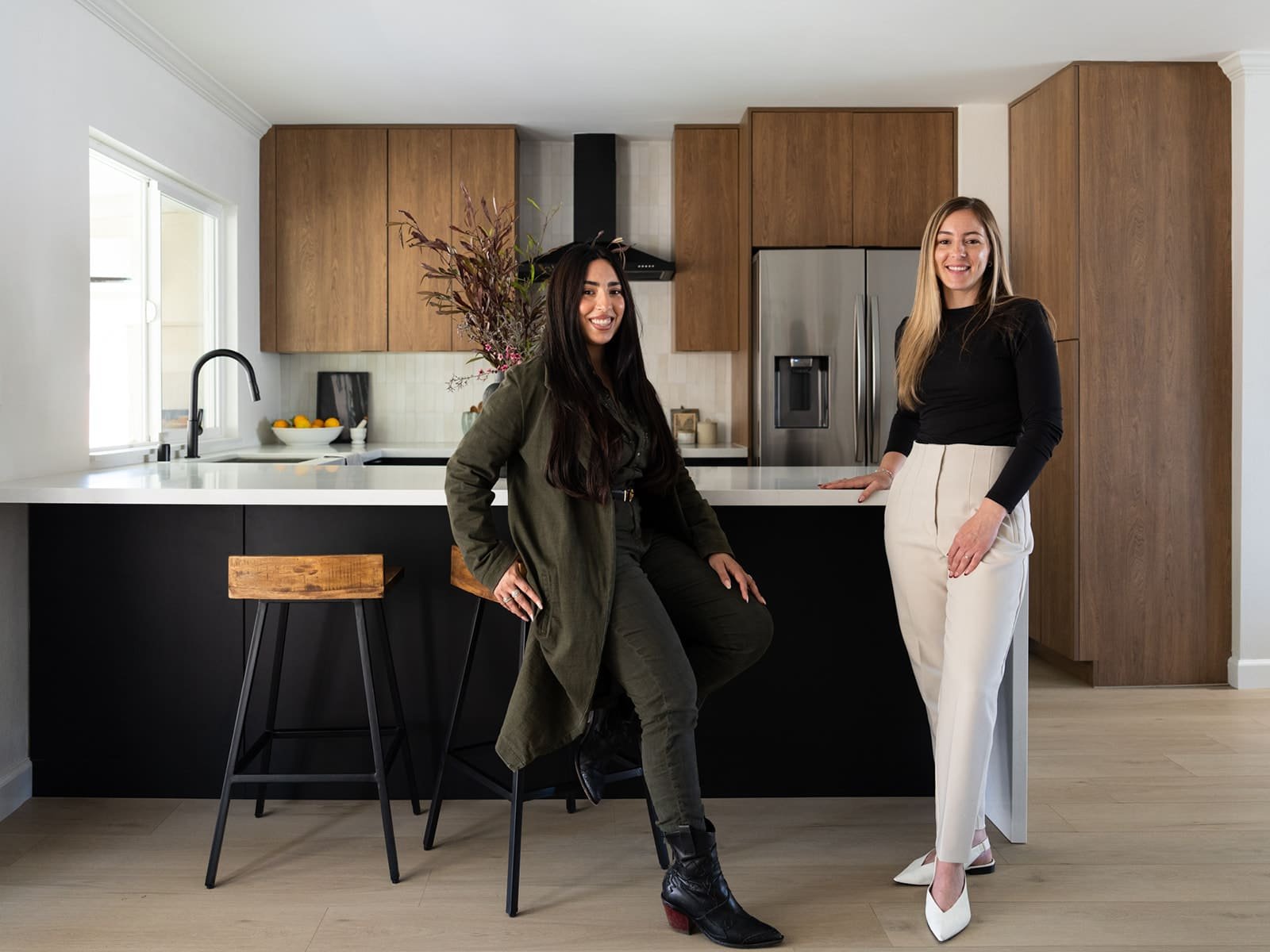 Two women in a modern kitchen with wooden cabinets and a center island. The woman on the left is sitting on a barstool, wearing a green outfit, and the woman on the right is standing, wearing a black top and white pants. There's a vase with branches