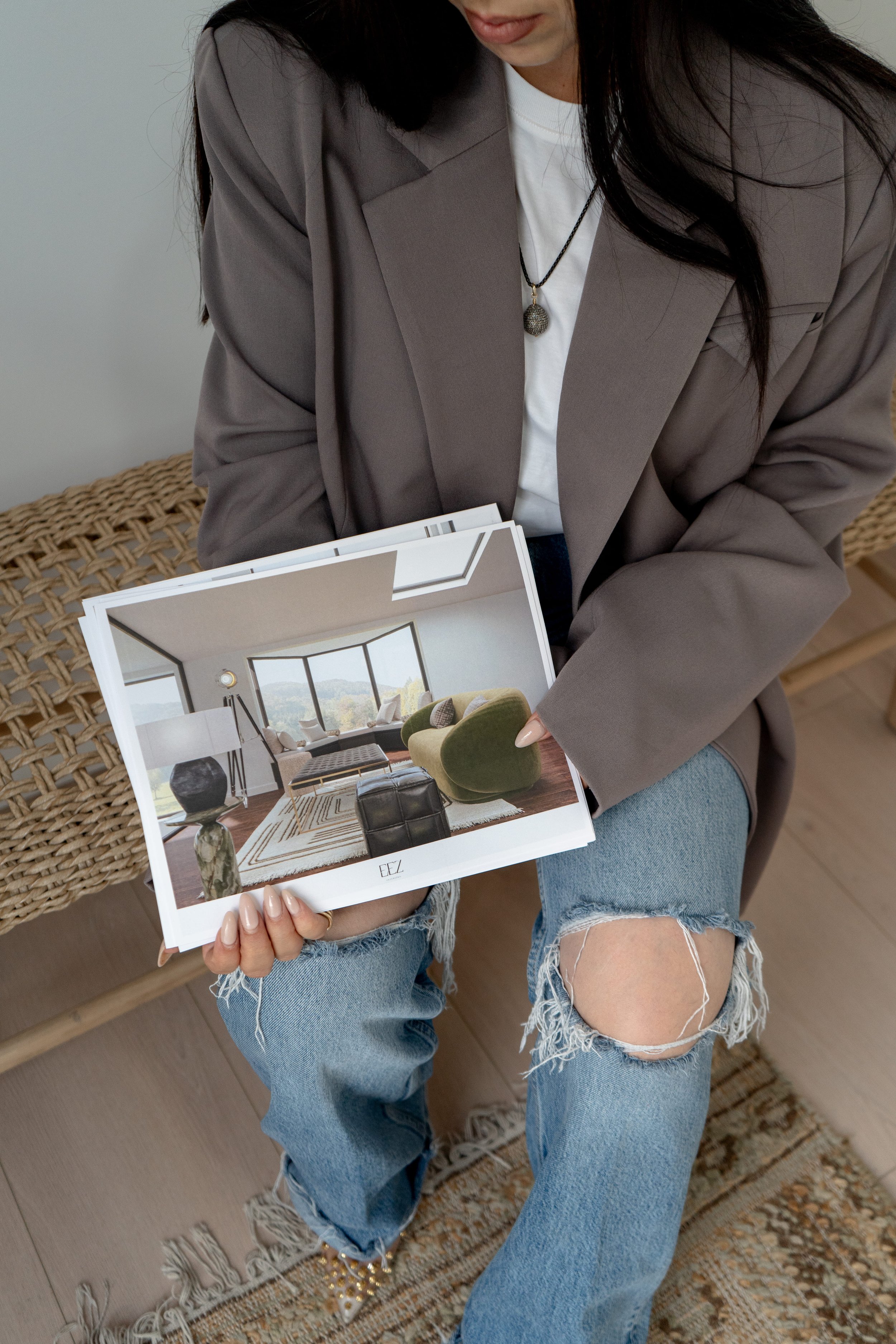 Person in a gray blazer holding an interior design magazine, showcasing a modern living room with green furniture, a large window, and a floor lamp. They're seated on a woven bench, wearing ripped jeans and a white shirt.