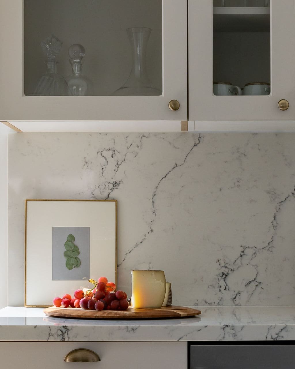 Marble kitchen countertop with a wooden cutting board holding red grapes and cheese, framed artwork leaning against the backsplash, glass cabinets above containing glassware.