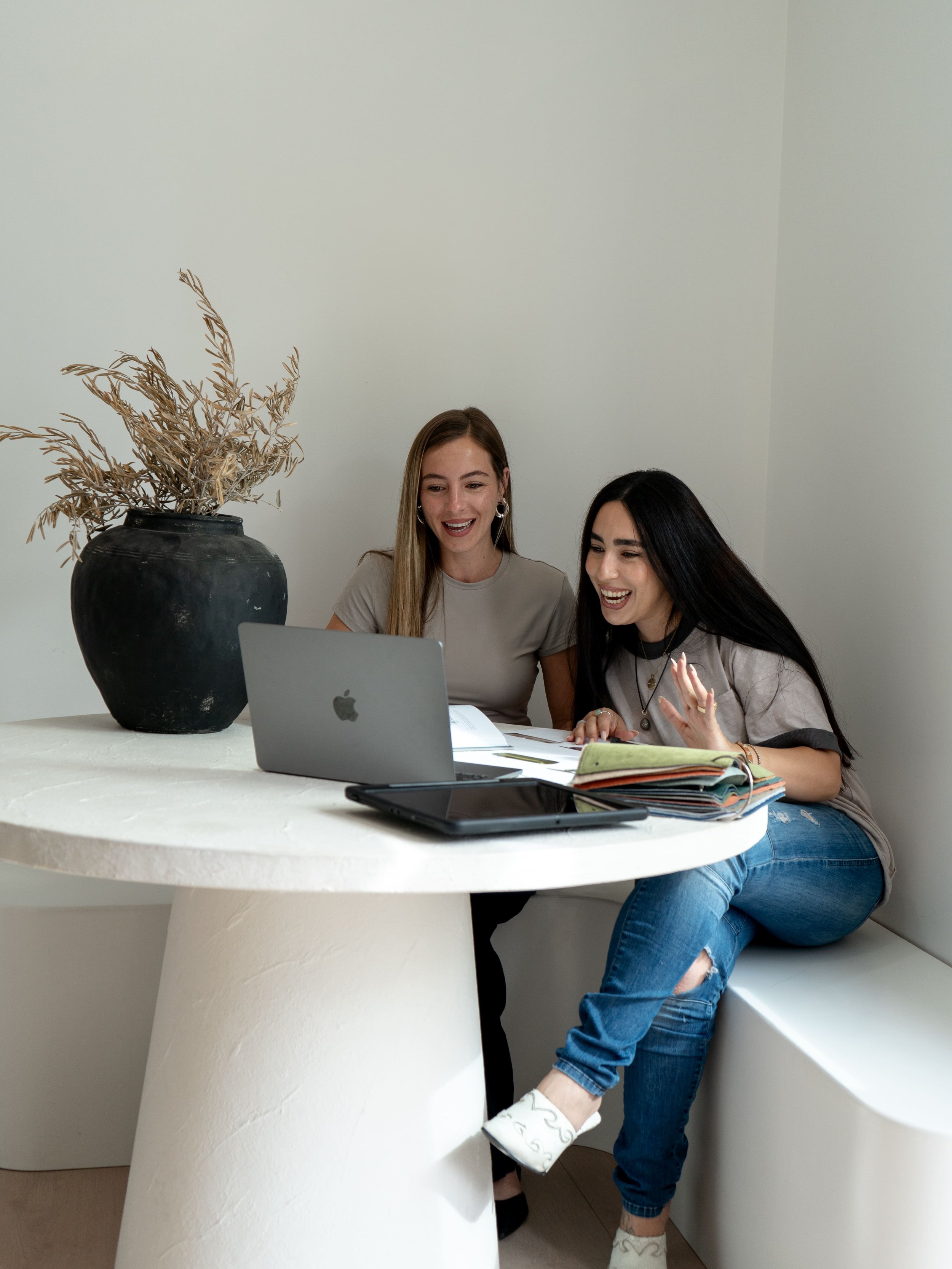 Two women sitting at a round white table, looking at a laptop, smiling. A large black vase with dried flowers is on the table. An open notebook and a tablet are also present.