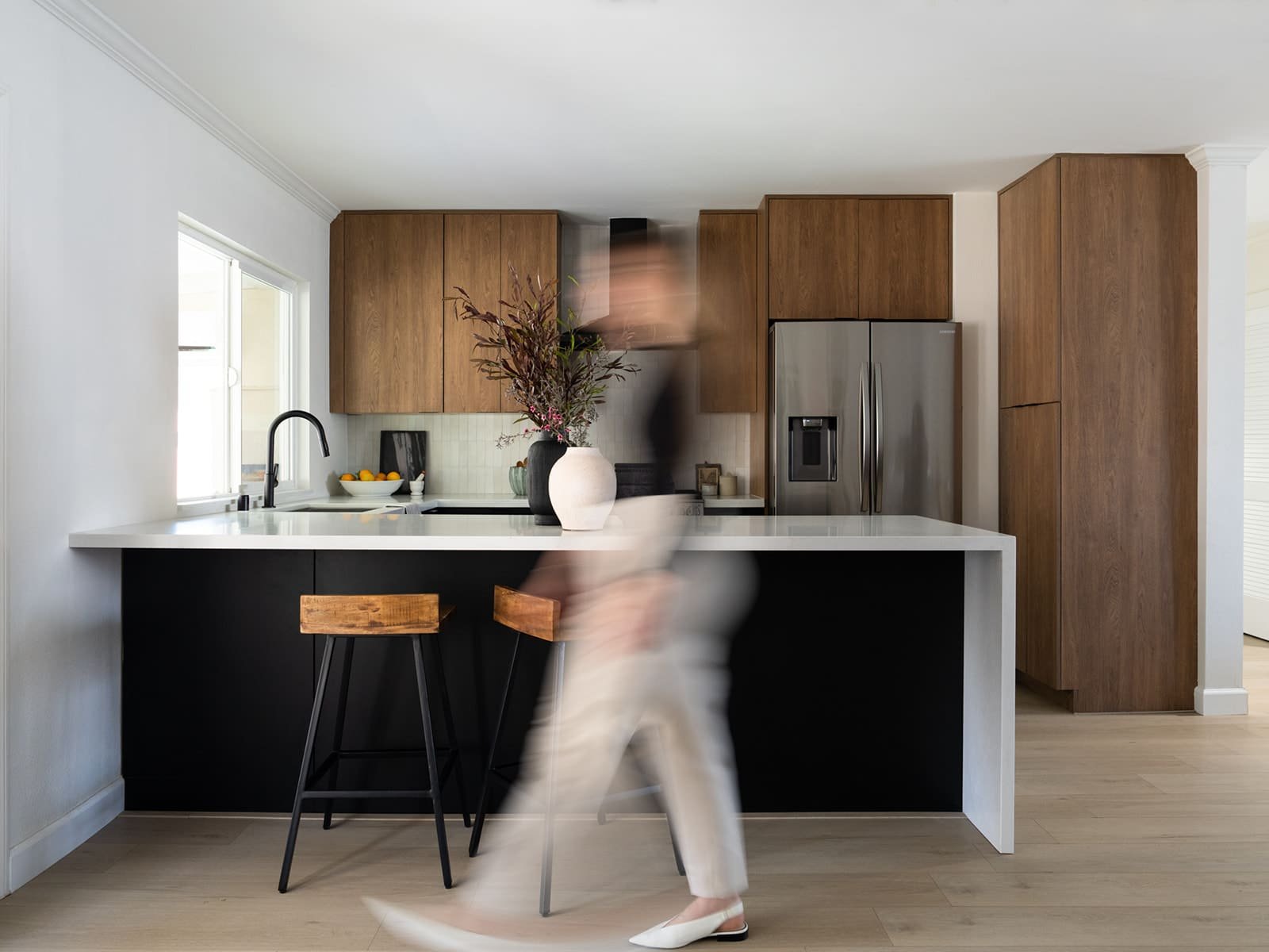 Modern kitchen with wooden cabinets, white countertop, stainless steel refrigerator, black sink, and two rustic stools. A vase with branches sits on the counter. A blurred figure is moving past the island.