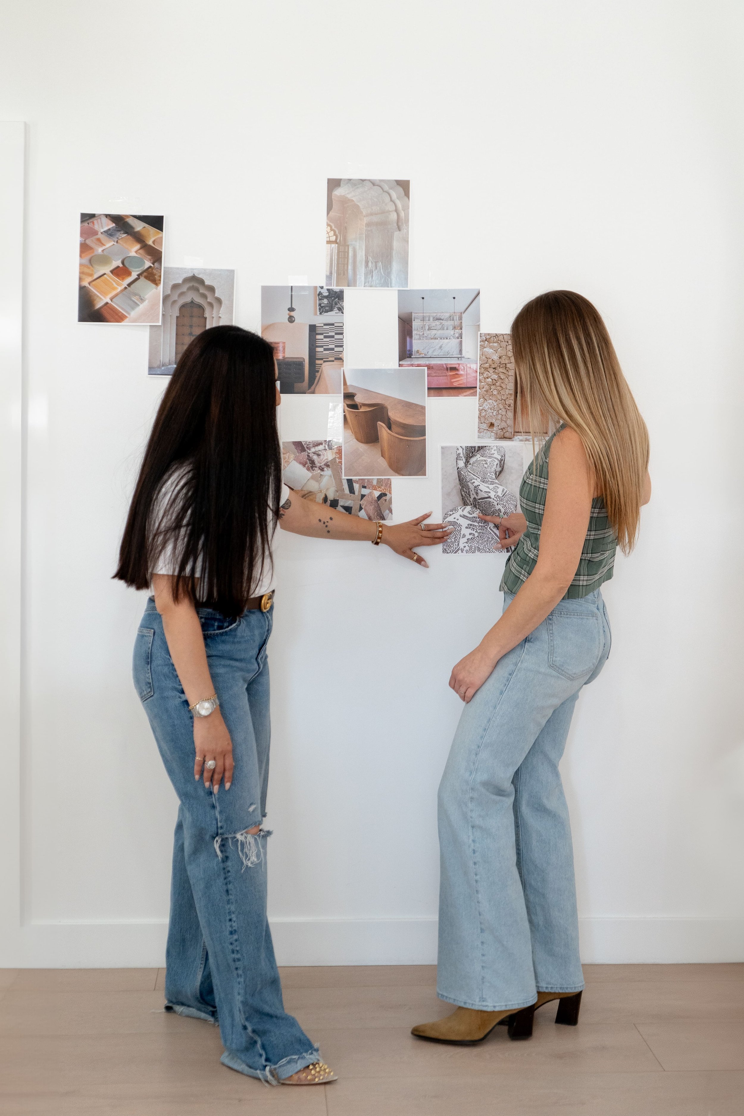 Two women in casual outfits stand in front of a wall with various architectural and interior design photos.