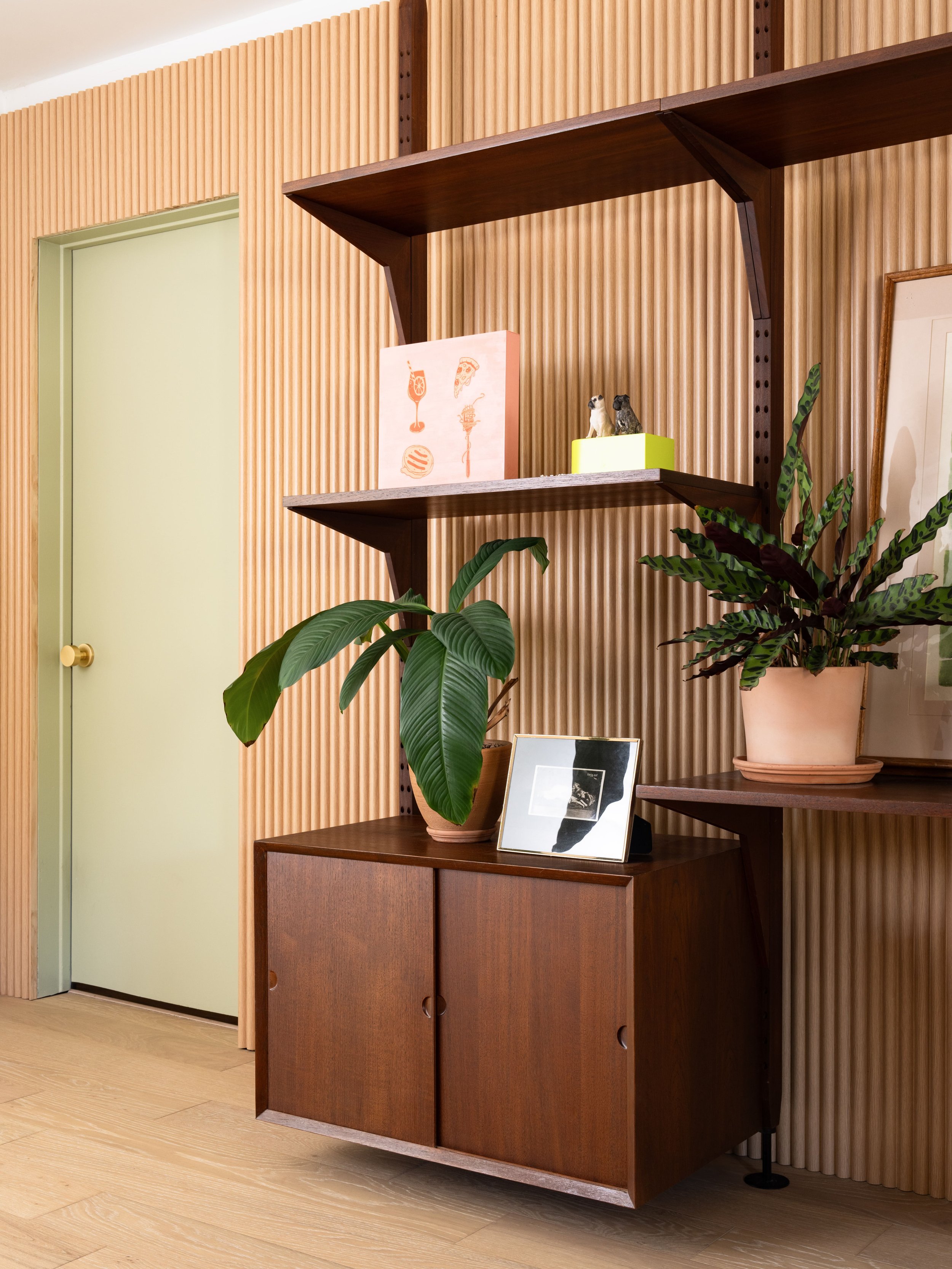 Modern interior with wooden shelves and cabinet against vertically grooved wall panels. Decor includes potted plants, a small framed picture, and artwork on the shelves. Light wood flooring and a closed light green door visible on the left.