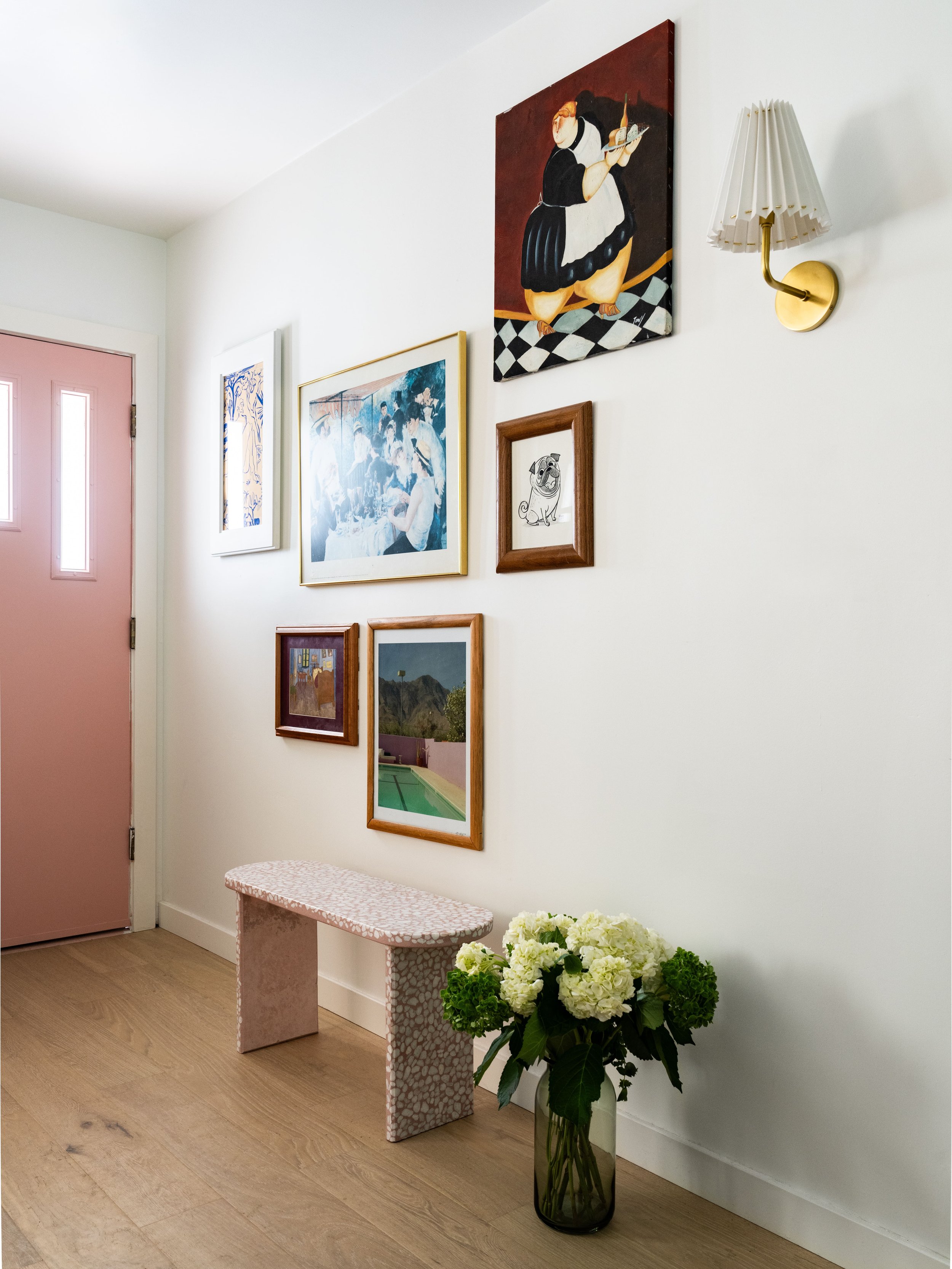 Entryway with pink door, wall art, small marble bench, and vase of white hydrangeas.