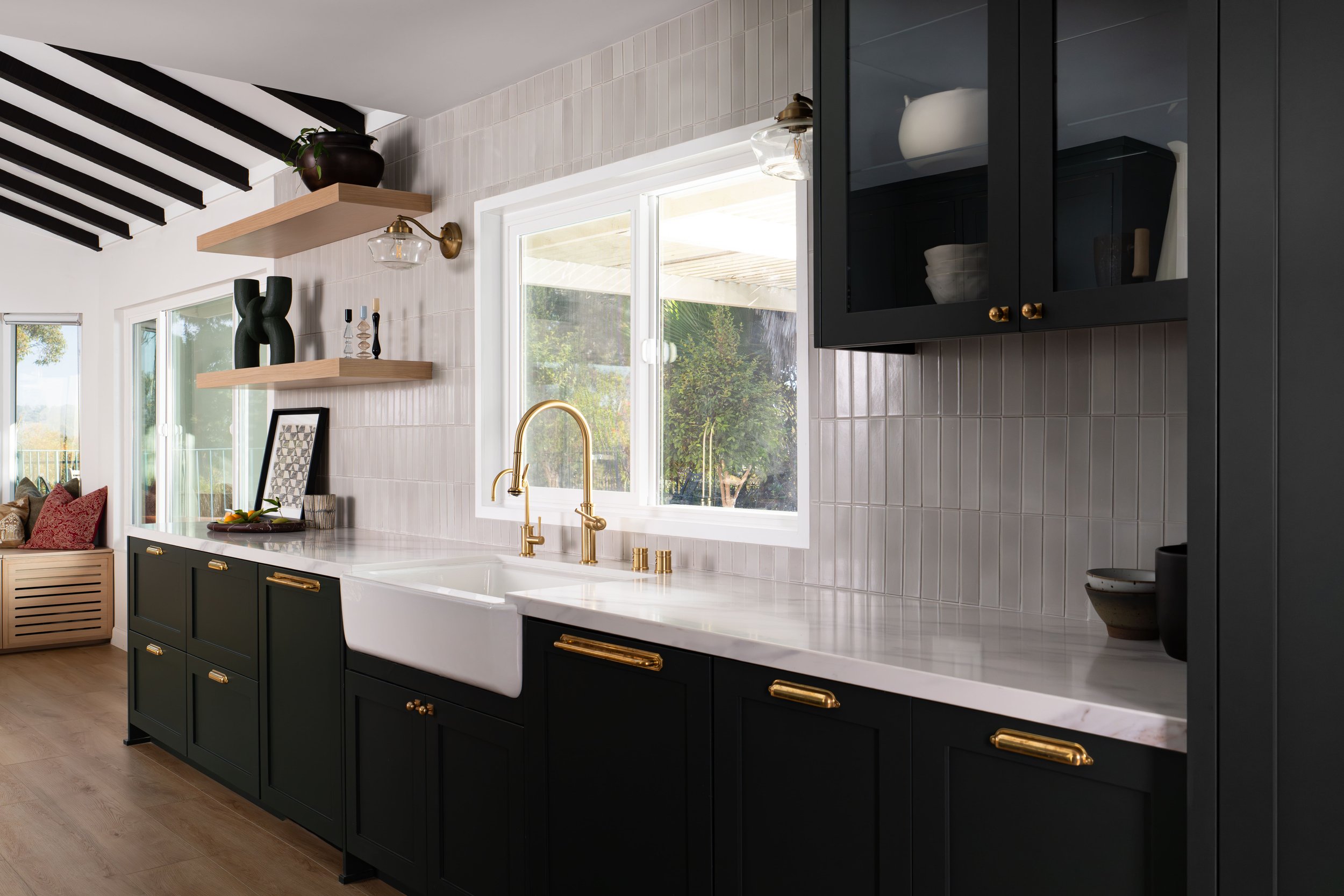 Modern kitchen interior with black cabinets, white countertops, and gold fixtures. Shelves with decorative items, a window with outdoor view, and a farmhouse sink.