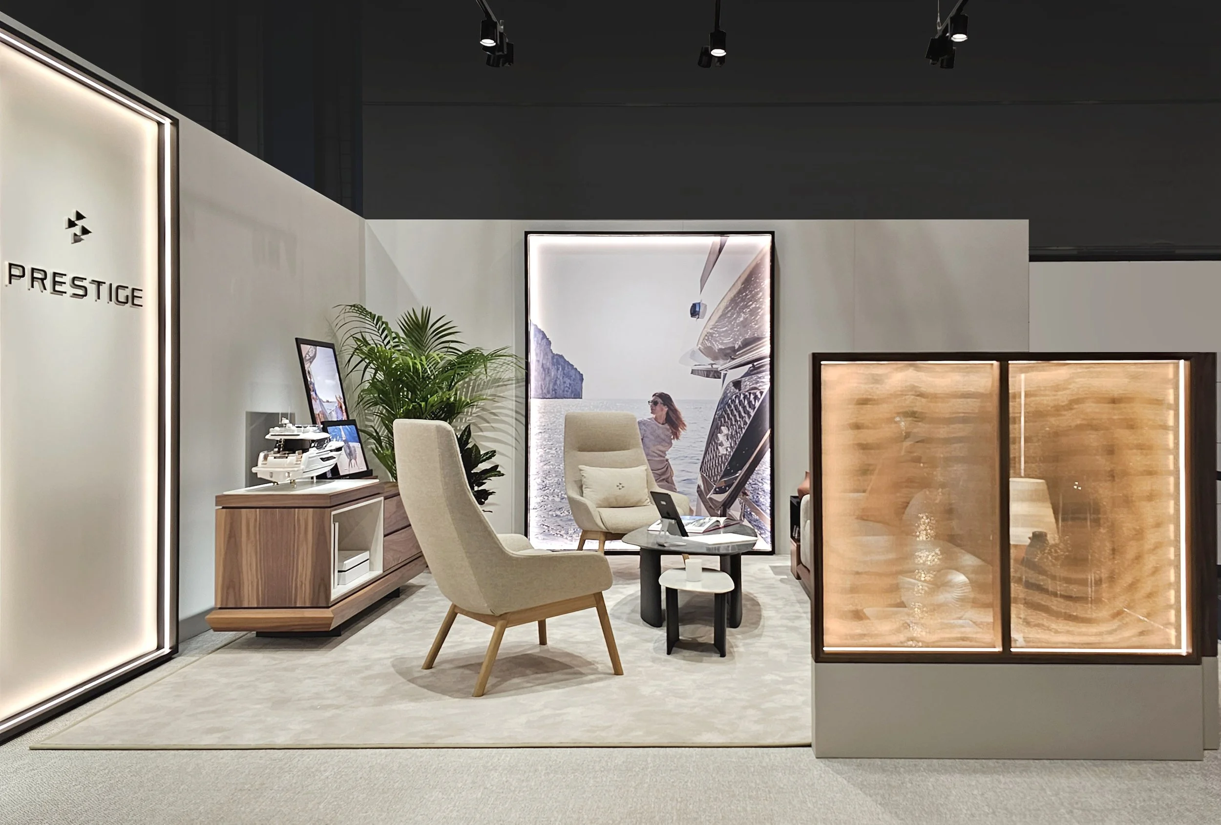 Une pièce de salon moderne avec un grand poster de paysage marin au fond, des meubles en bois clair, des fauteuils beige, une plante verte, et un mur lumineux avec le logo Prestige.