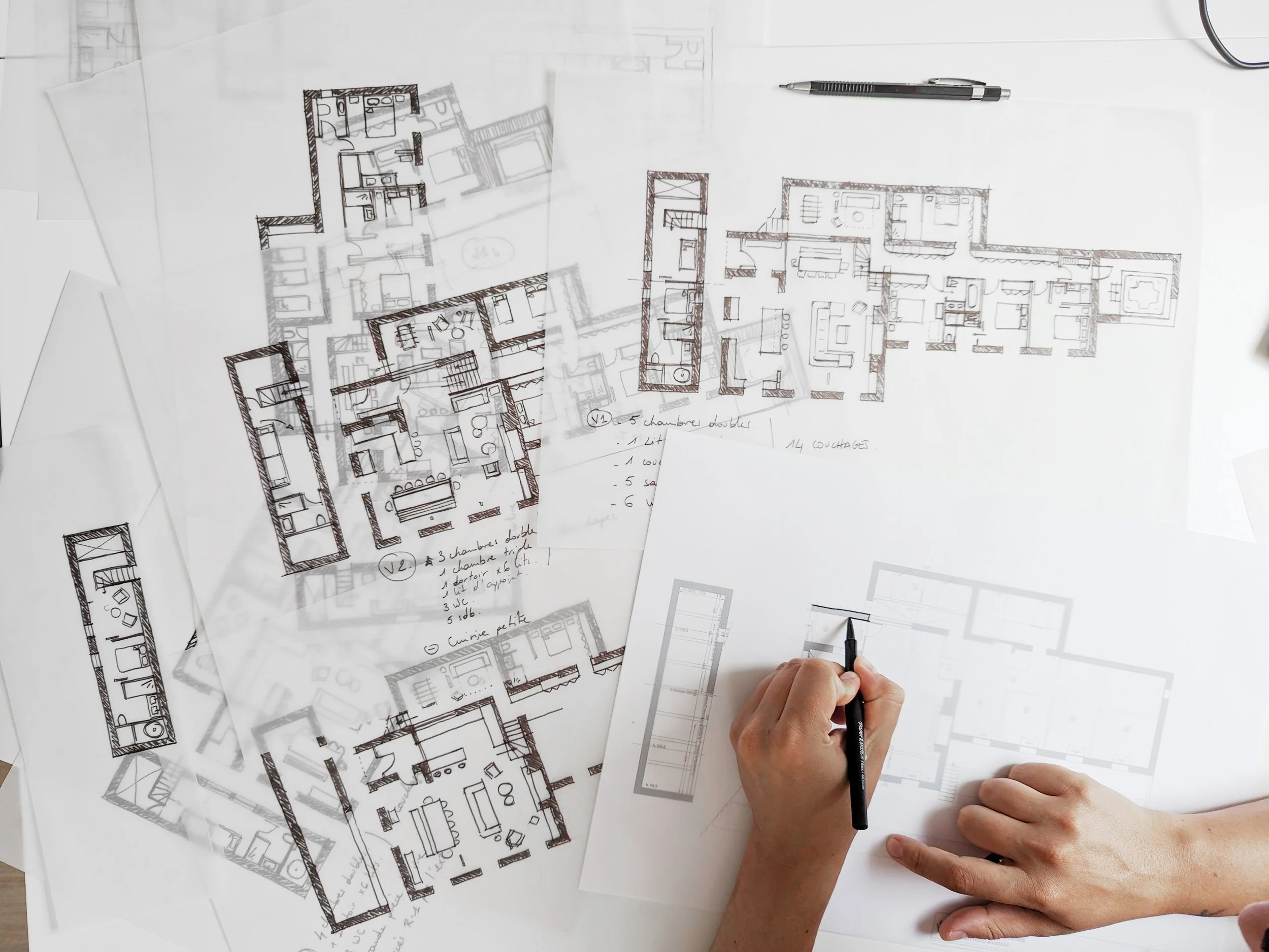 Des plans architecturaux sur une table, avec une personne en train de dessiner et de prendre des notes. Un stylo est visible à côté des mains.