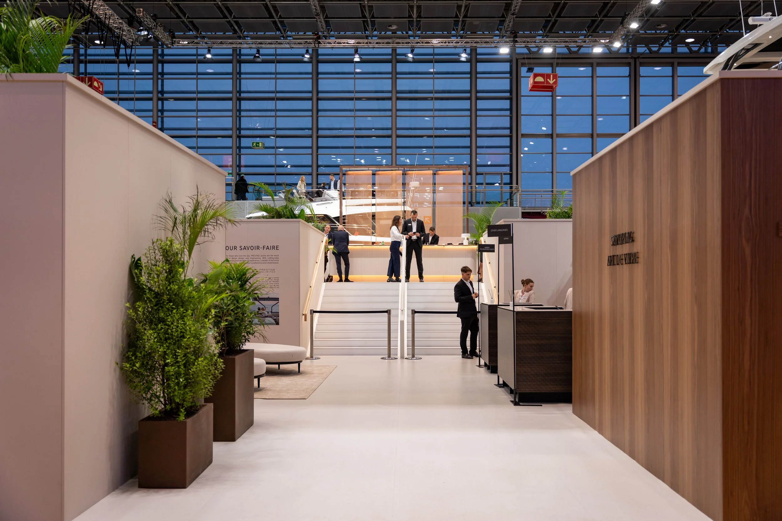 Vue intérieure d'un hall d'exposition ou de réception avec des plantes en pot, une zone d'accueil, des escaliers menant à un niveau supérieur, et des personnes en train de discuter ou d'utiliser leurs téléphones, sous une grande verrière.