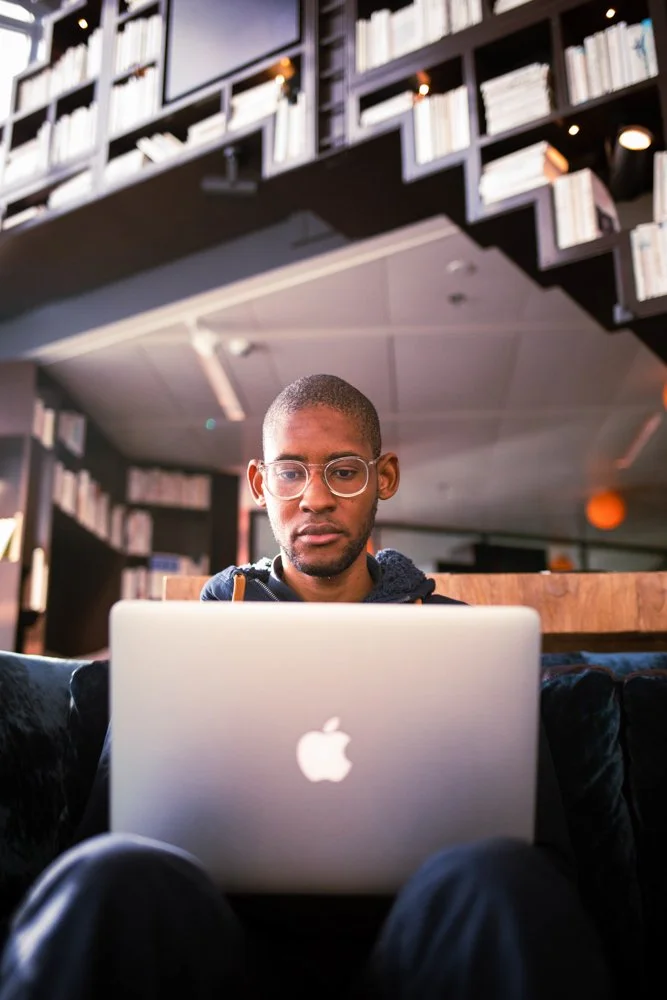 Homme assis devant un ordinateur portable Apple dans un environnement intérieur, probablement une bibliothèque ou un café, avec des étagères pleines de livres en arrière-plan.