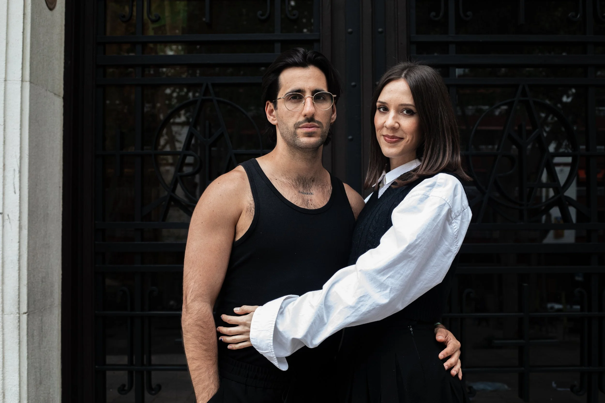 Un homme et une femme se tiennent côte à côte devant une grille noire. L'homme porte un débardeur noir et porte des lunettes rondes, la femme porte une chemise blanche et un pull noir. Elle a les bras autour de l'homme.