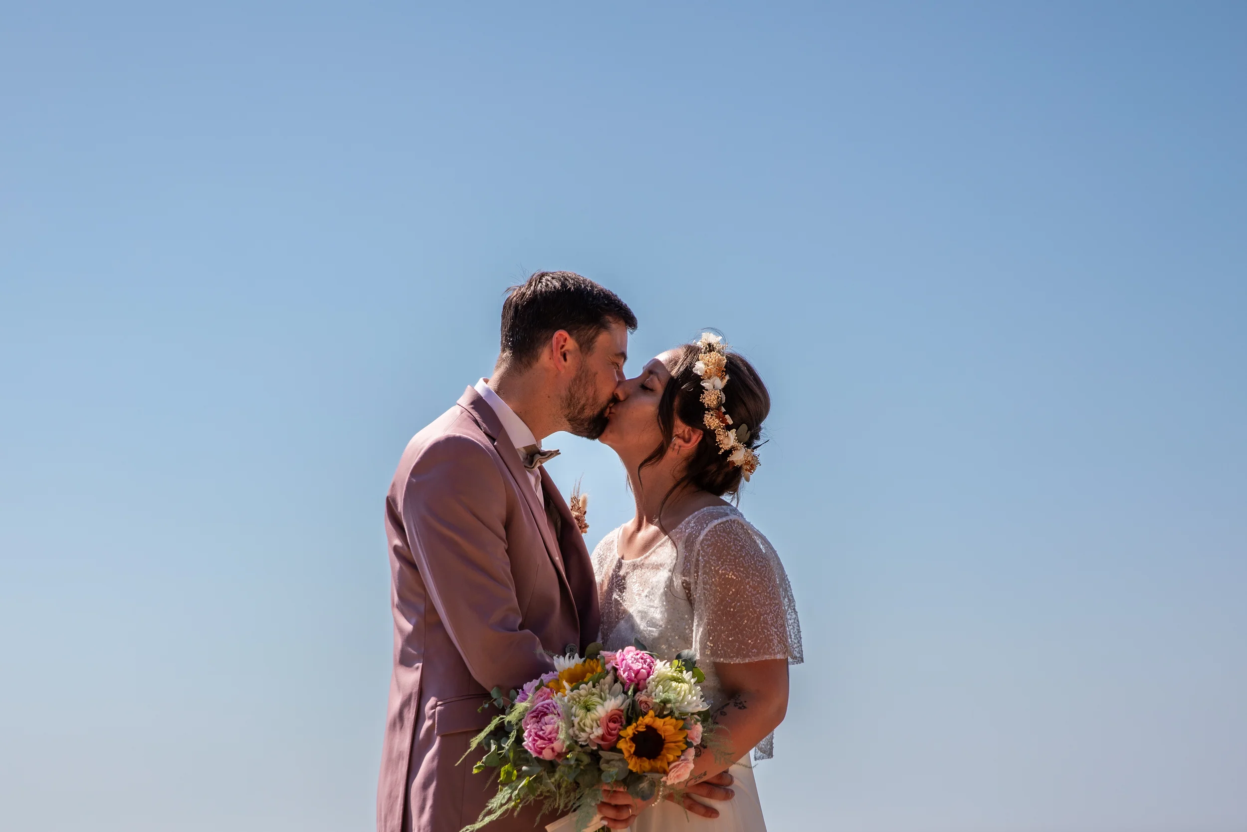 Un couple en mariage s'embrassant sous un ciel bleu clair, la femme porte une couronne de fleurs et tient un bouquet coloré.