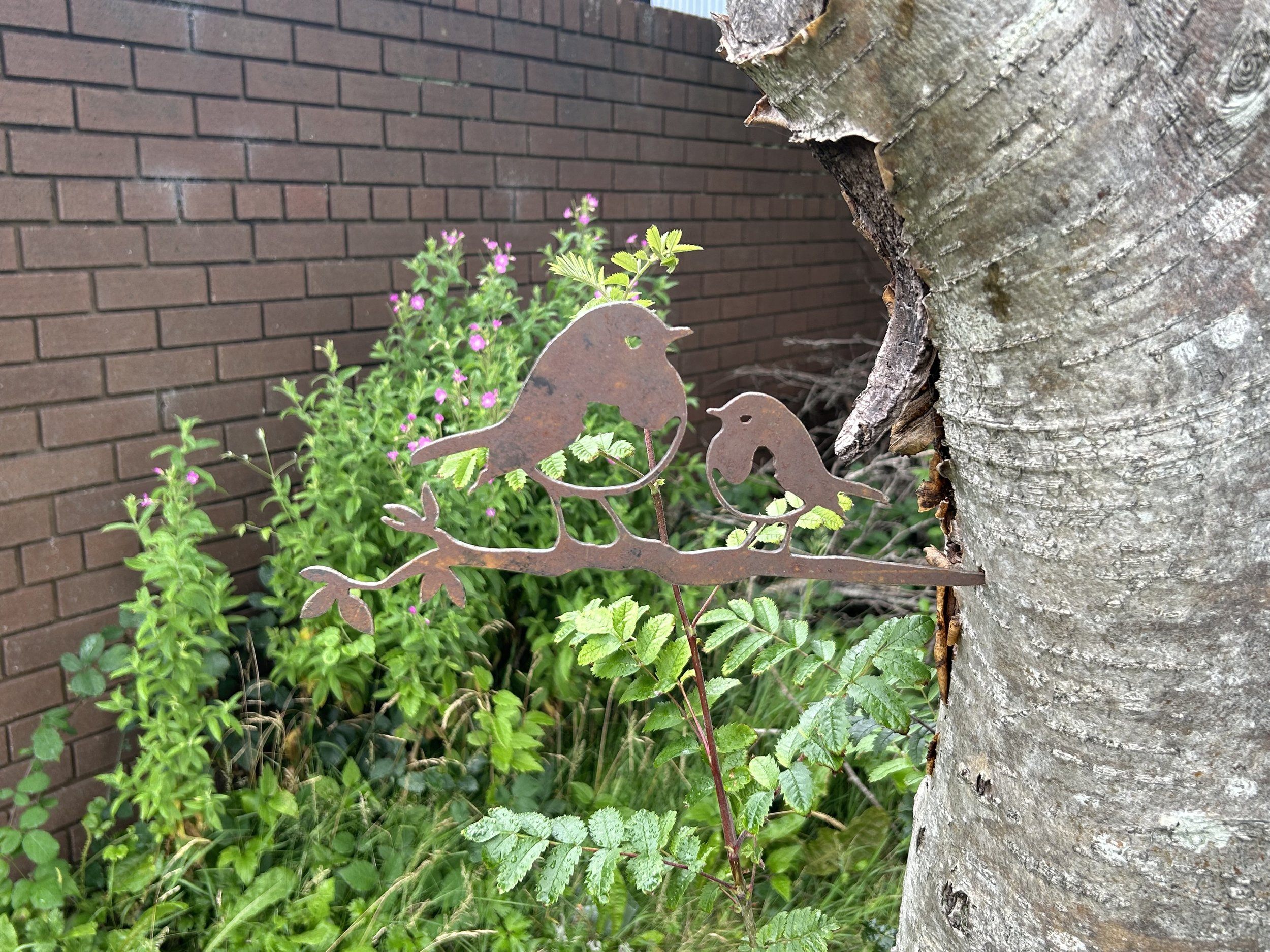 Metal robin ornament attached to a tree, with young green leaves and tall wildflowers growing behind it against a brown brick wall