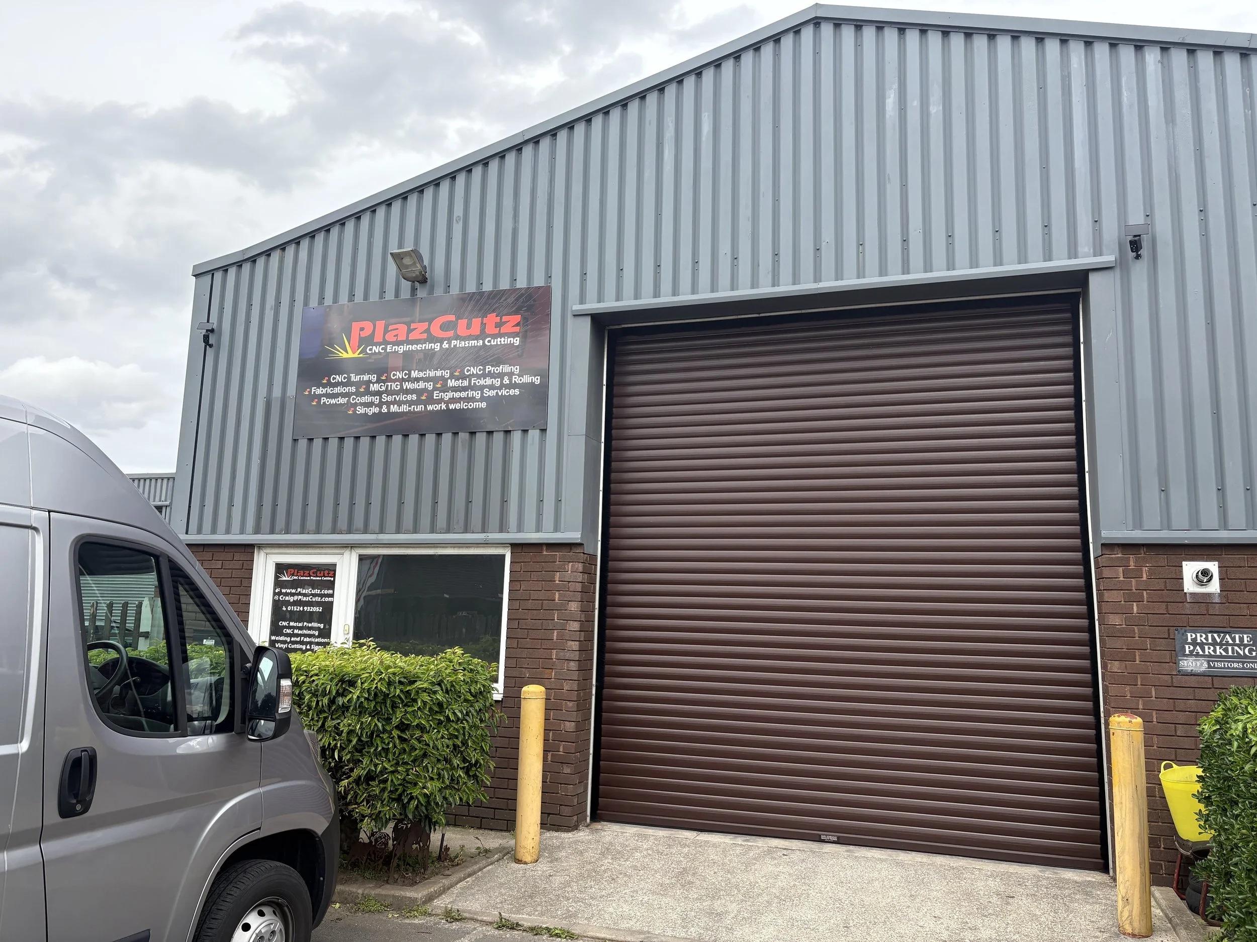 Business building with a sign that reads 'PlazCutz CNC Engineering & Plasma Cutting,' featuring a list of services including CNC turning, CNC machining, fabrication, MIG/TIG welding, metal folding, rolling, powder coating, engineering services, and welding. There is a roller shutter door, a security camera, a silver van parked in front, and a sign indicating a private parking area.