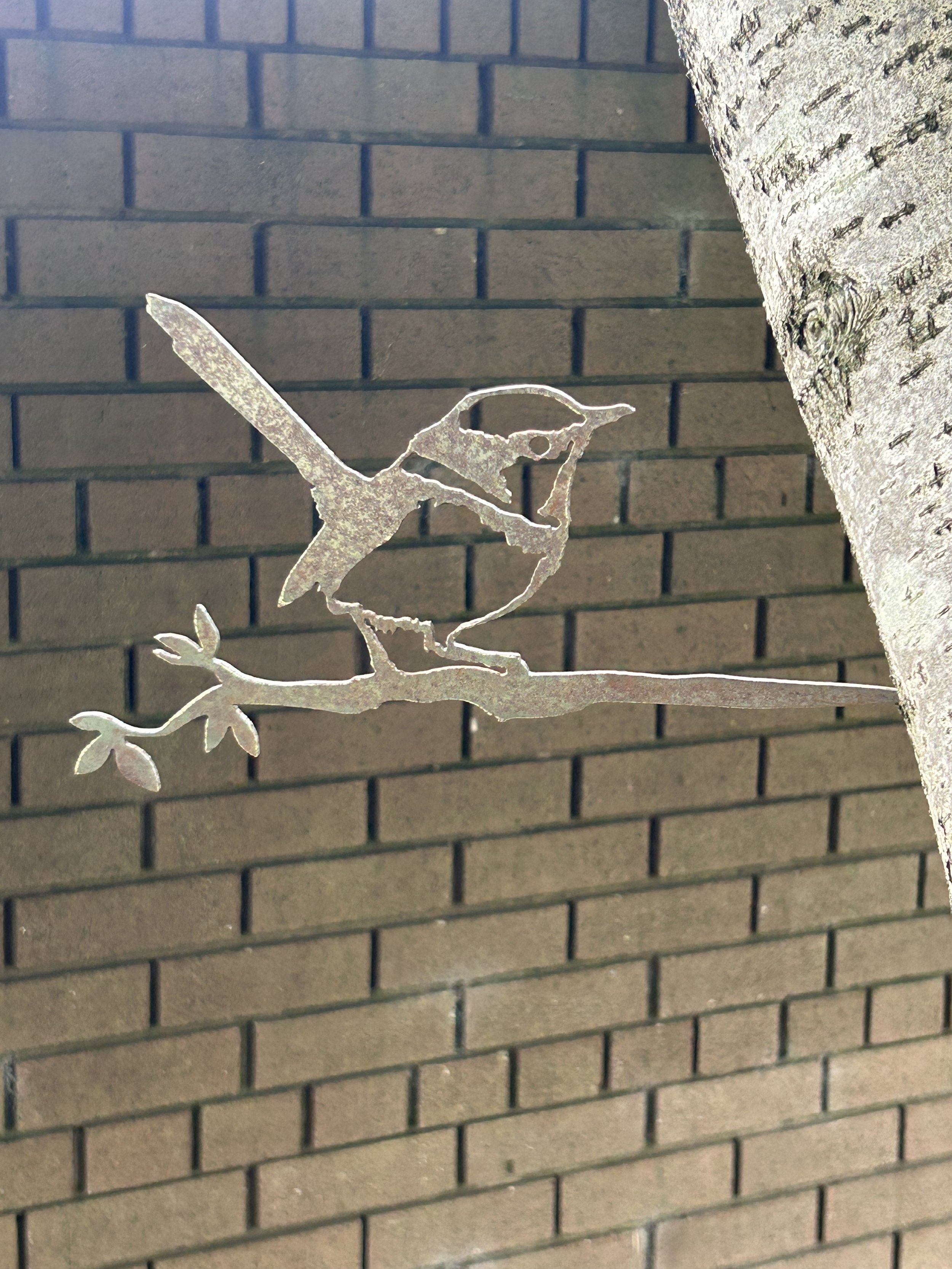 Rustic metalbird silhouette of a Wren perched on a branch, attached to a tree trunk with a brown brick wall in the background, creating a contrast between nature and urban texture