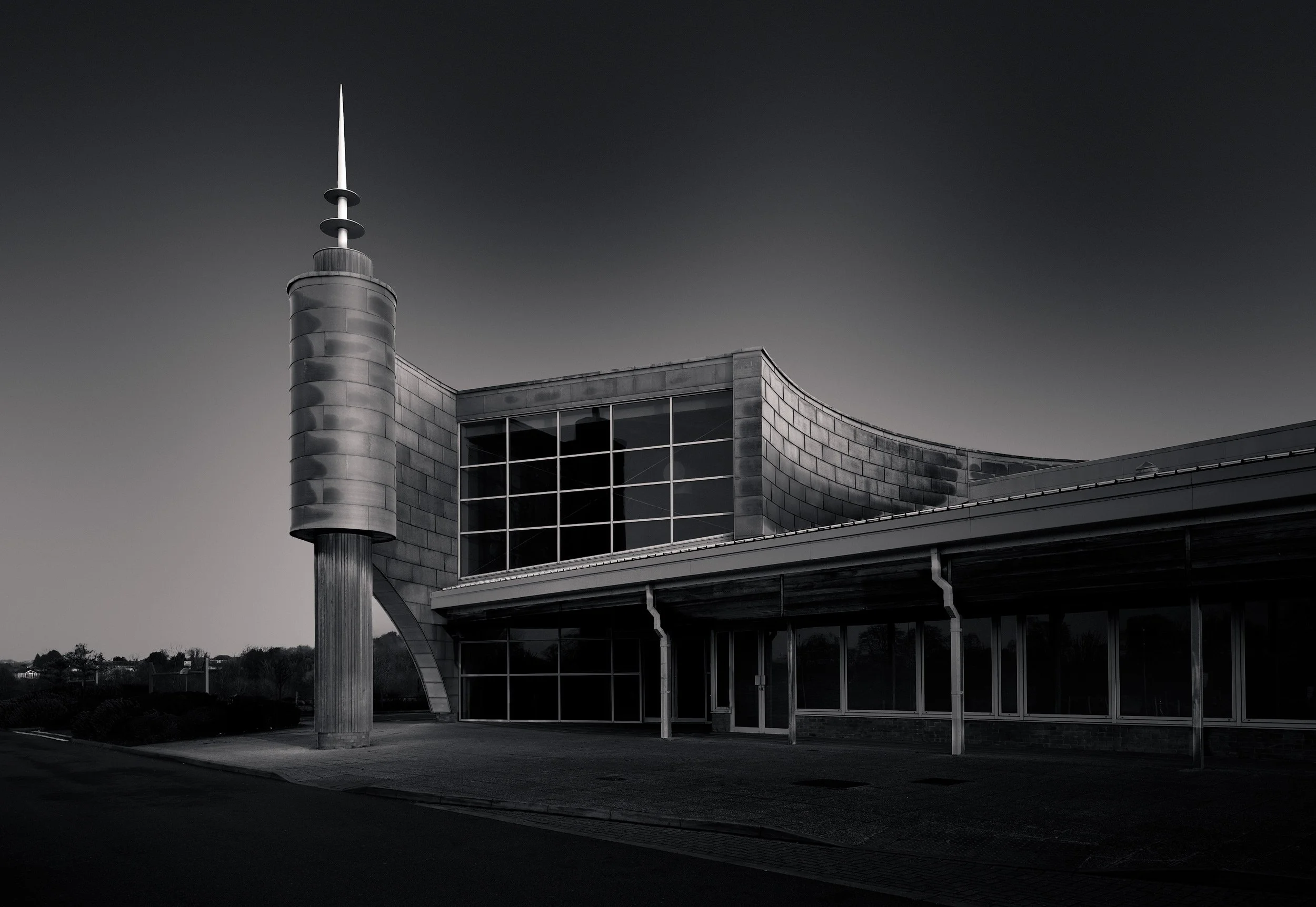 [architecture] [uk] [evesham] [blackandwhite] Evesham Leisure Centre zinc clad tower with spire, curved façade and glazed frontage, moody black and white monochrome. Architectural Photography by Jim Scott.