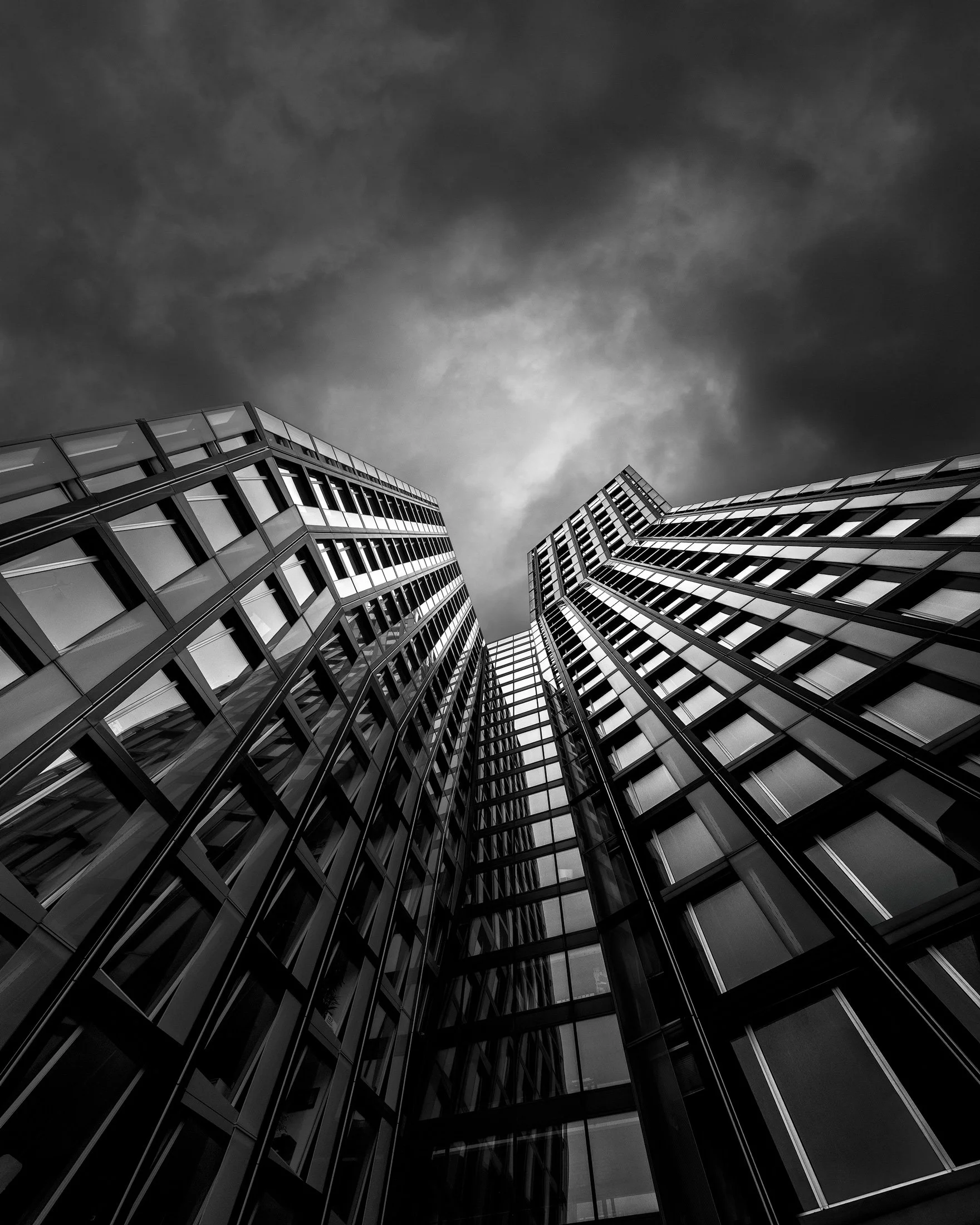 Modern skyscrapers in Hamburg: The Dancing Towers (Tanzende Türme) on the Reeperbahn. A dramatic low-angle black and white architectural photograph of the 'bending' glass towers against a stormy, dark sky.