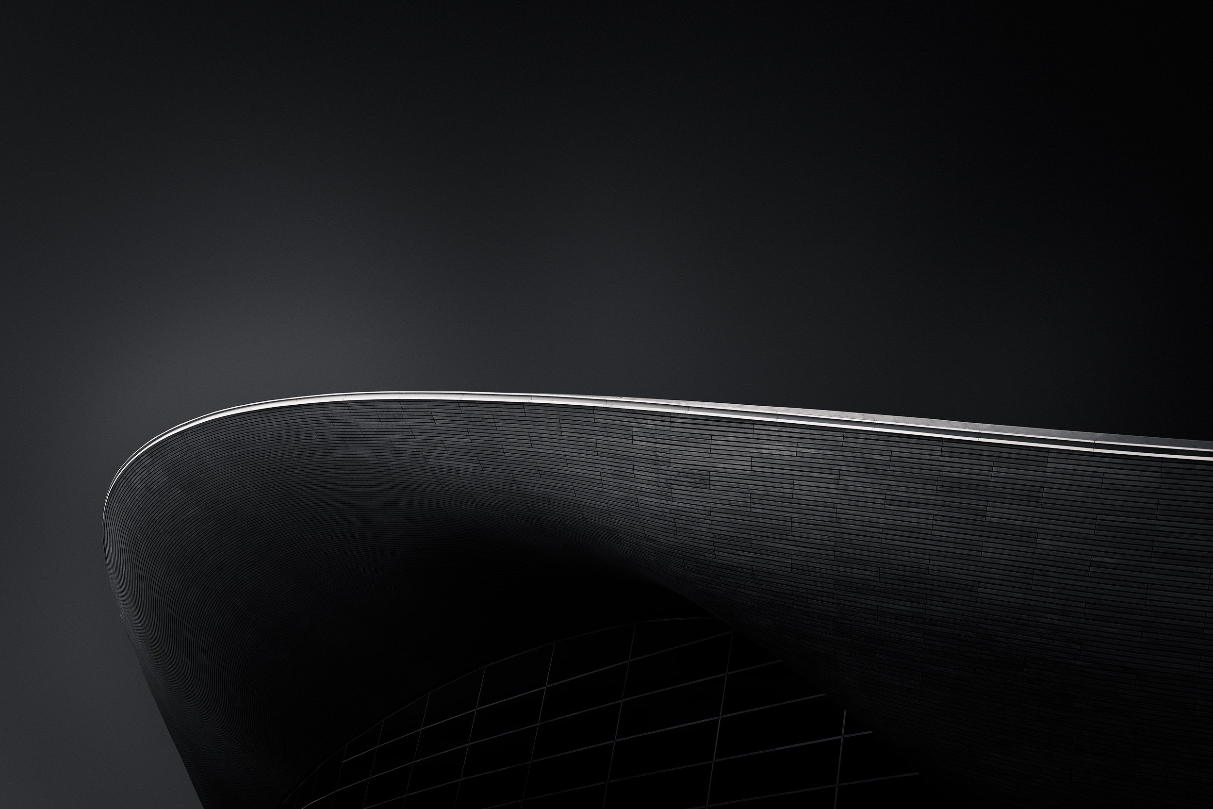 Parametric architecture in Stratford, London: A minimalist monochromatic study of the London Aquatics Centre, designed by Zaha Hadid Architects. This architectural photograph captures the fluid, wave-inspired roofline isolated against a dark, minimal