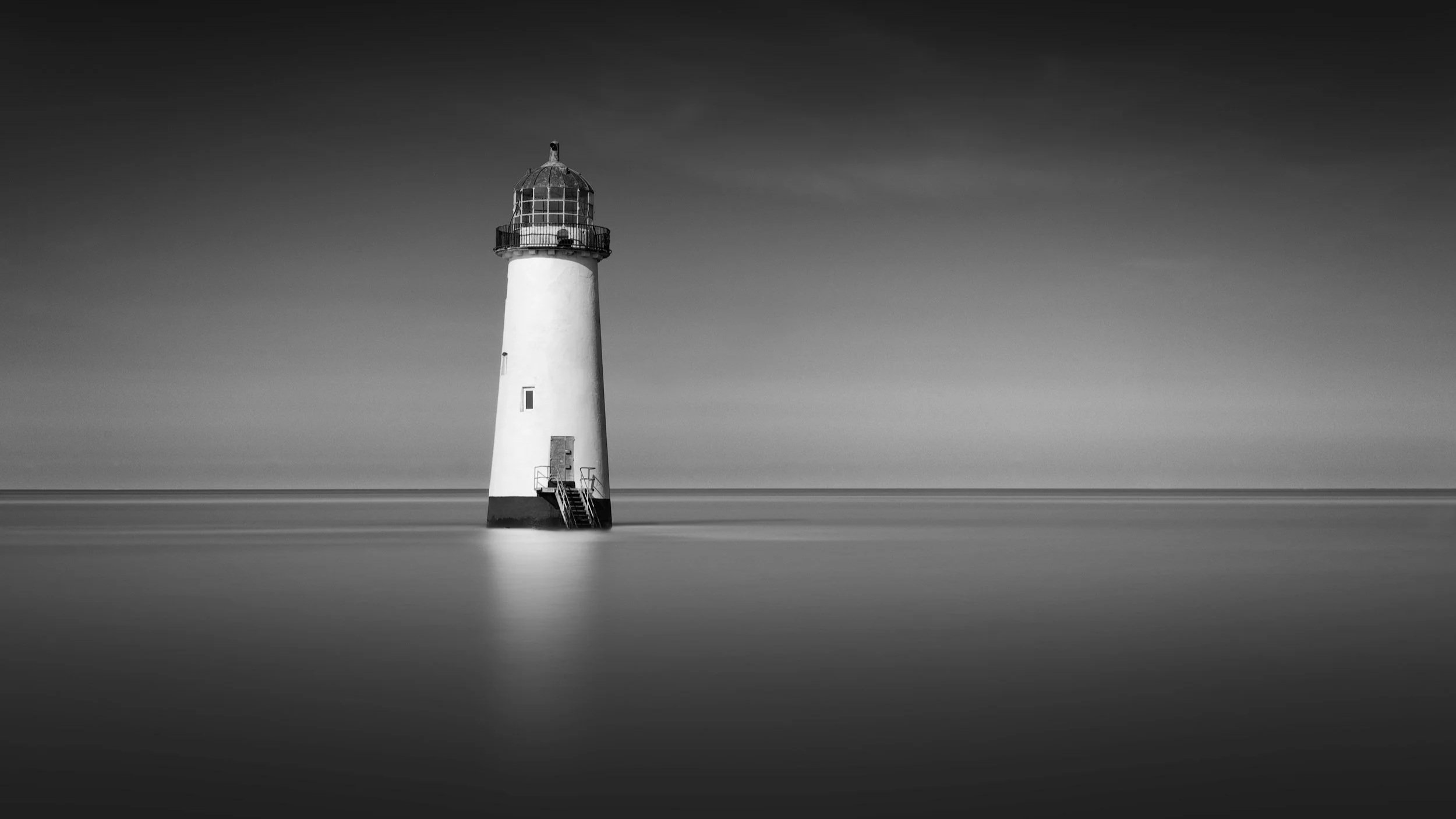 [architecture] [uk] [talacre] [blackandwhite] [panoramic] Talacre Lighthouse Wales long exposure B&W, white cylindrical tower standing alone on a dark base above a clam sea. Architectural Photography by Jim Scott.