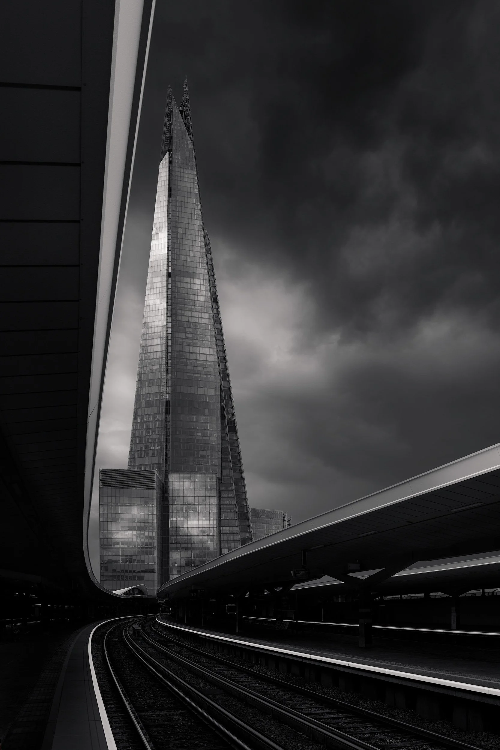 [architecture] [uk] [london] [blackandwhite] The Shard from London Bridge station, tapering glass spire rising behind curving platform canopies under a storm sky. Architectural Photography by Jim Scott.