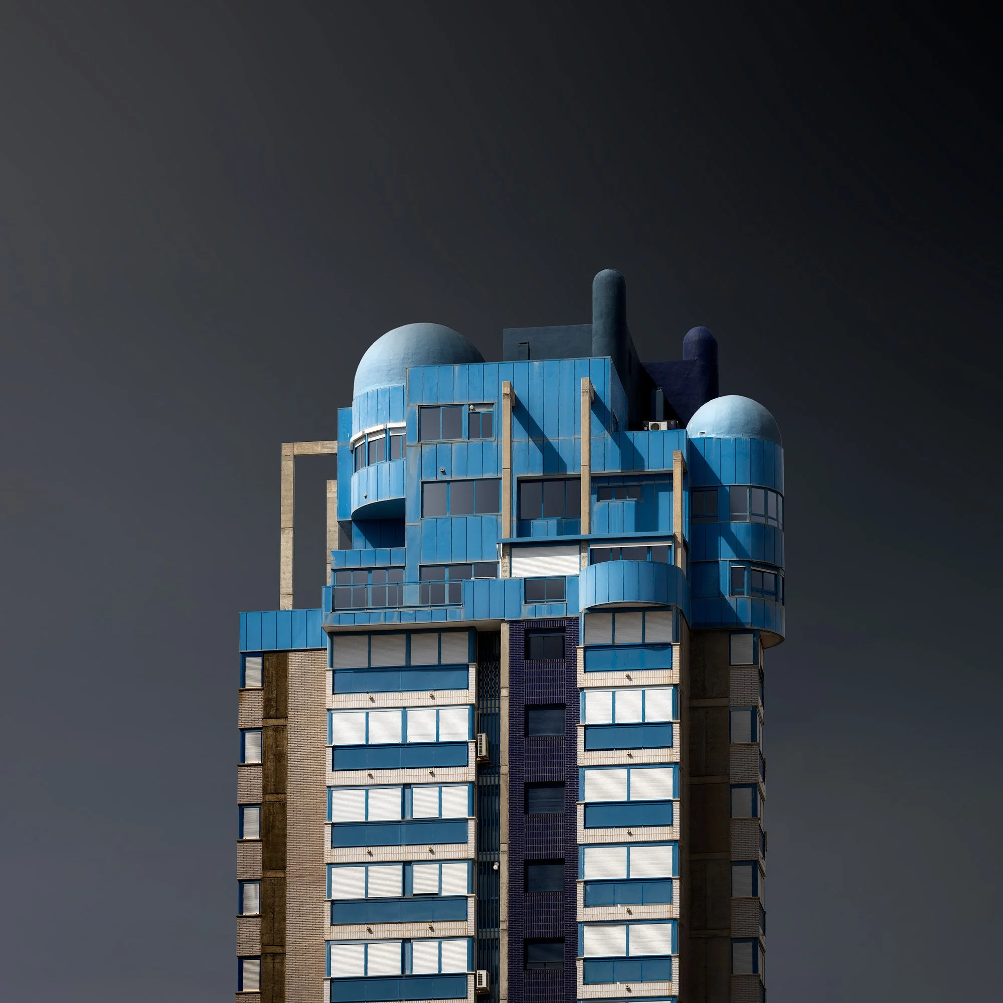 [architecture] [colour] [europe] [benidorm] [square] Edificio Kennedy III Benidorm, blue-panelled façade crowned with dome structures against a dark sky. Architectural Photography by Jim Scott.