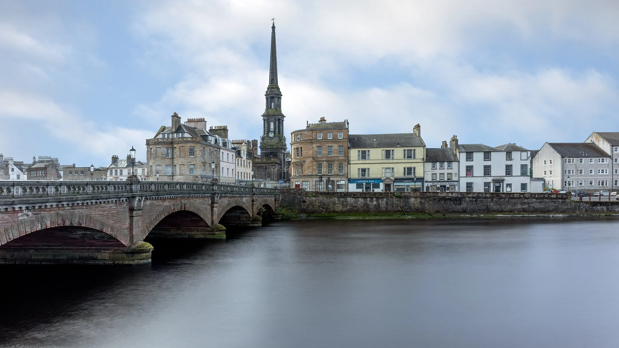 Traditional Scottish urbanism in Ayr town centre: A serene long-exposure study of the historic New Bridge of Ayr spanning the River Ayr. This landscape photograph captures the warm sandstone architecture and the prominent spire of the Ayr Town Hall a