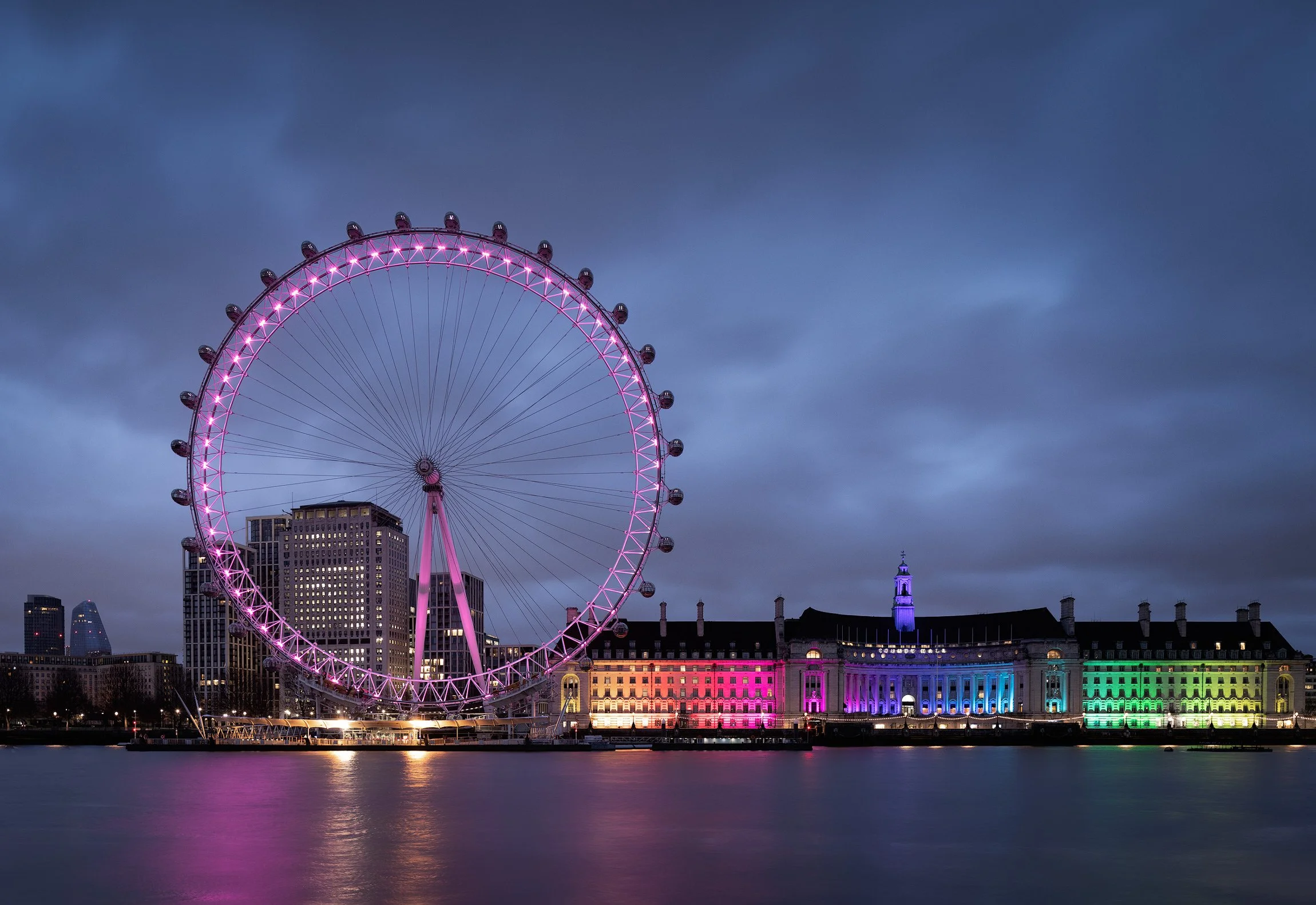 [architecture] [uk] [london] [colour] London Eye glowing pink at night, County Hall in rainbow colours beside the dark reflective River Thames. Architectural Photography by Jim Scott.