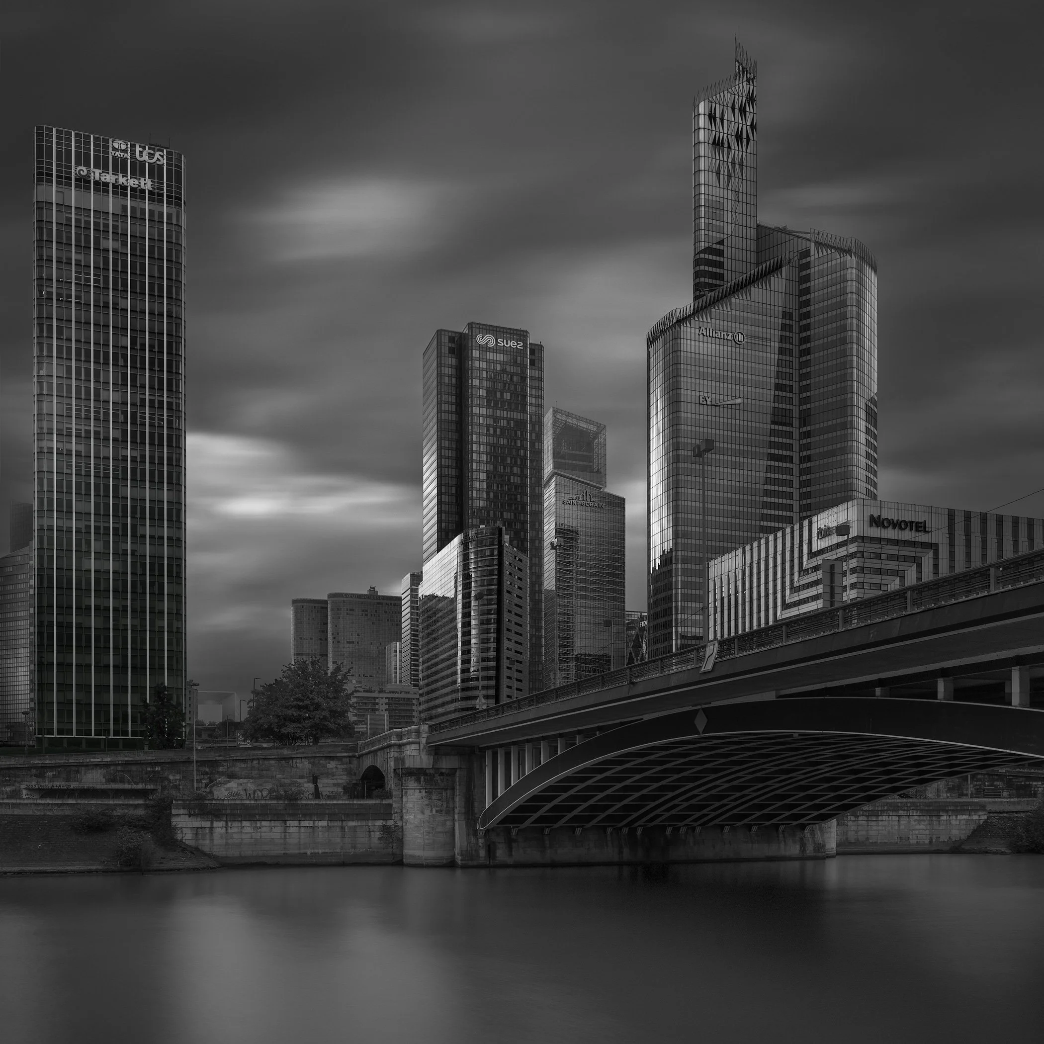 Contemporary commercial and infrastructure architecture in La Défense, Paris: A dramatic monochromatic study of the business district skyline from the Pont de Neuilly. This architectural photograph captures a dense cluster of glass-clad towers, inclu
