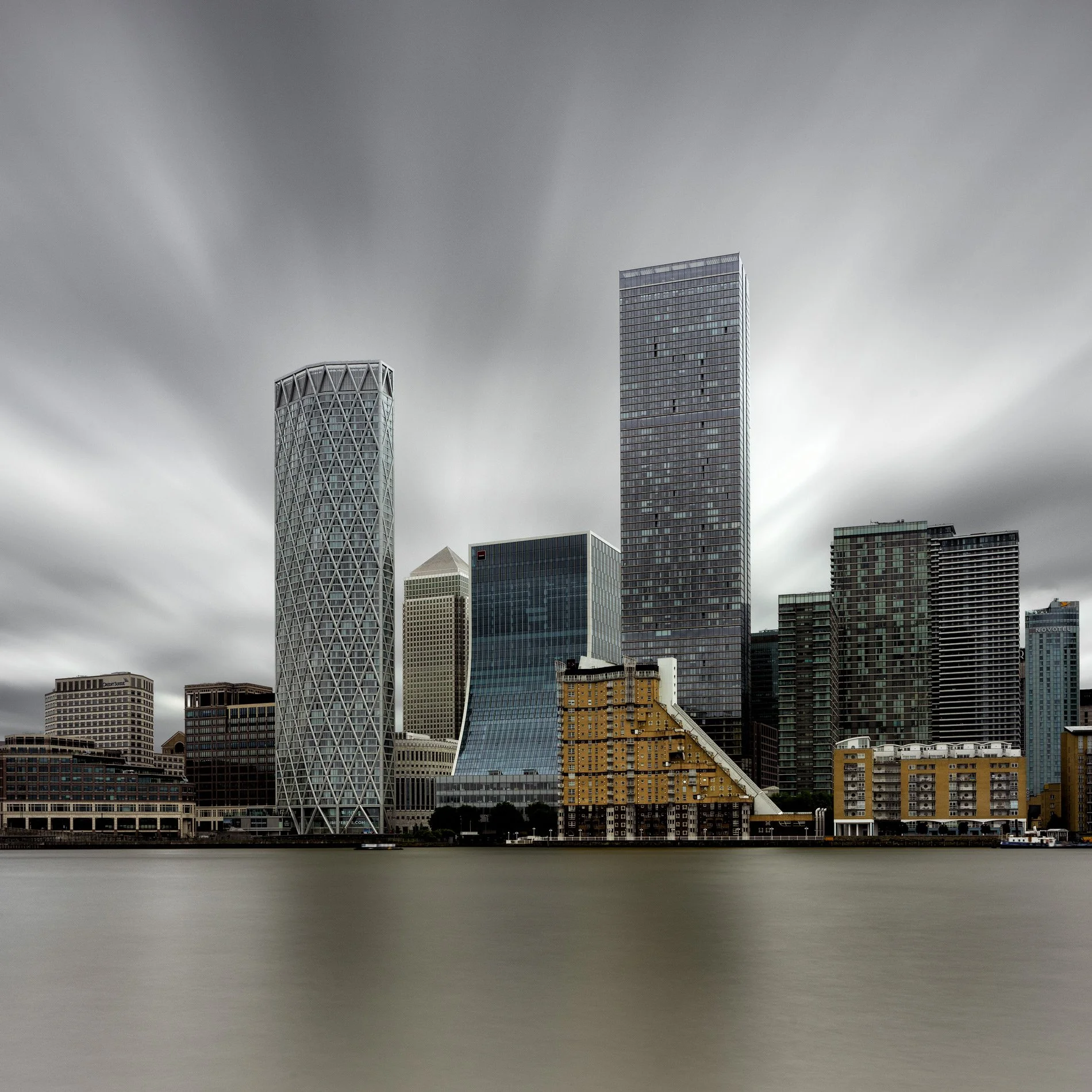 Modern skyscraper architecture in London: The Canary Wharf skyline featuring One Canada Square and Newfoundland tower. A long-exposure colour photograph across the River Thames, capturing the contrast between the cylindrical geometric towers and the 