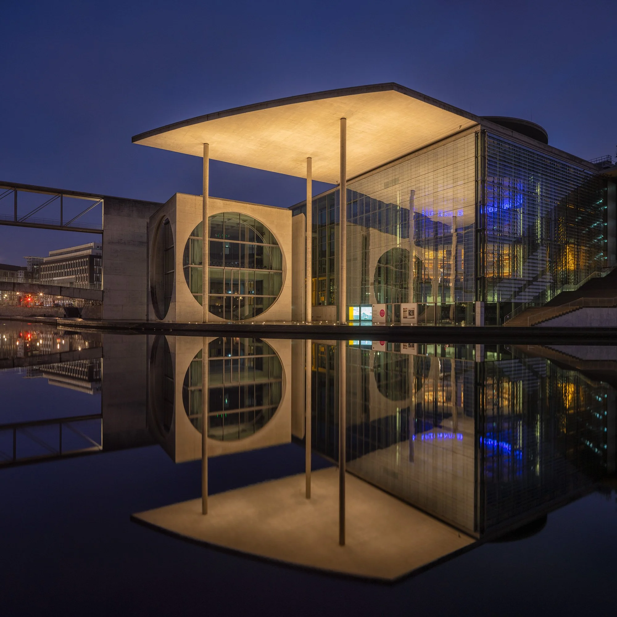 Contemporary civic architecture at twilight: A vibrant blue-hour study of the Marie-Elisabeth-Lüders-Haus in Berlin, designed by Stephan Braunfels. This symmetrical photograph captures the building’s iconic circular geometry and massive cantilevered 