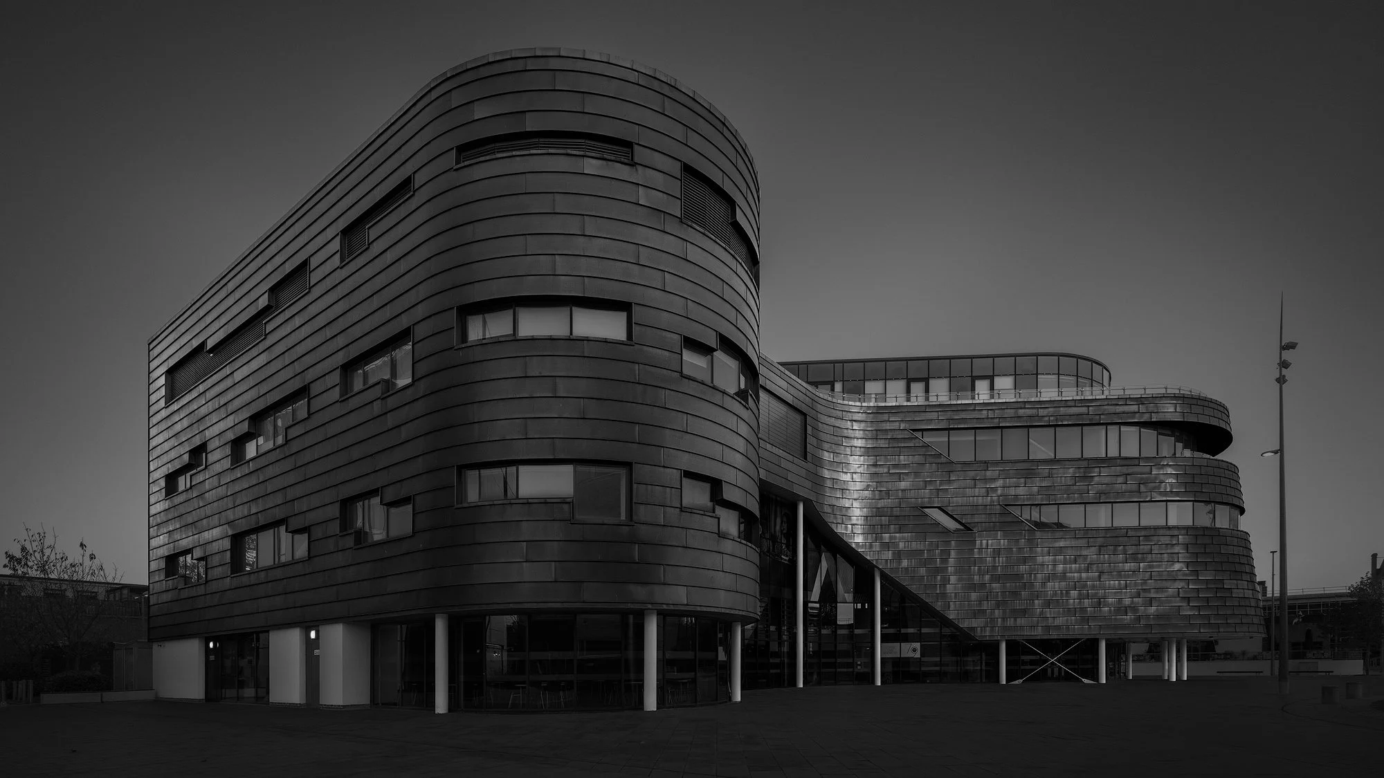 [architecture] [uk] [middlesbrough] [blackandwhite] [panoramic] Teesside University curved zinc clad facade raised on pilotis, sweeping horizontal banding, moody black and white monochrome. Architectural Photography by Jim Scott.