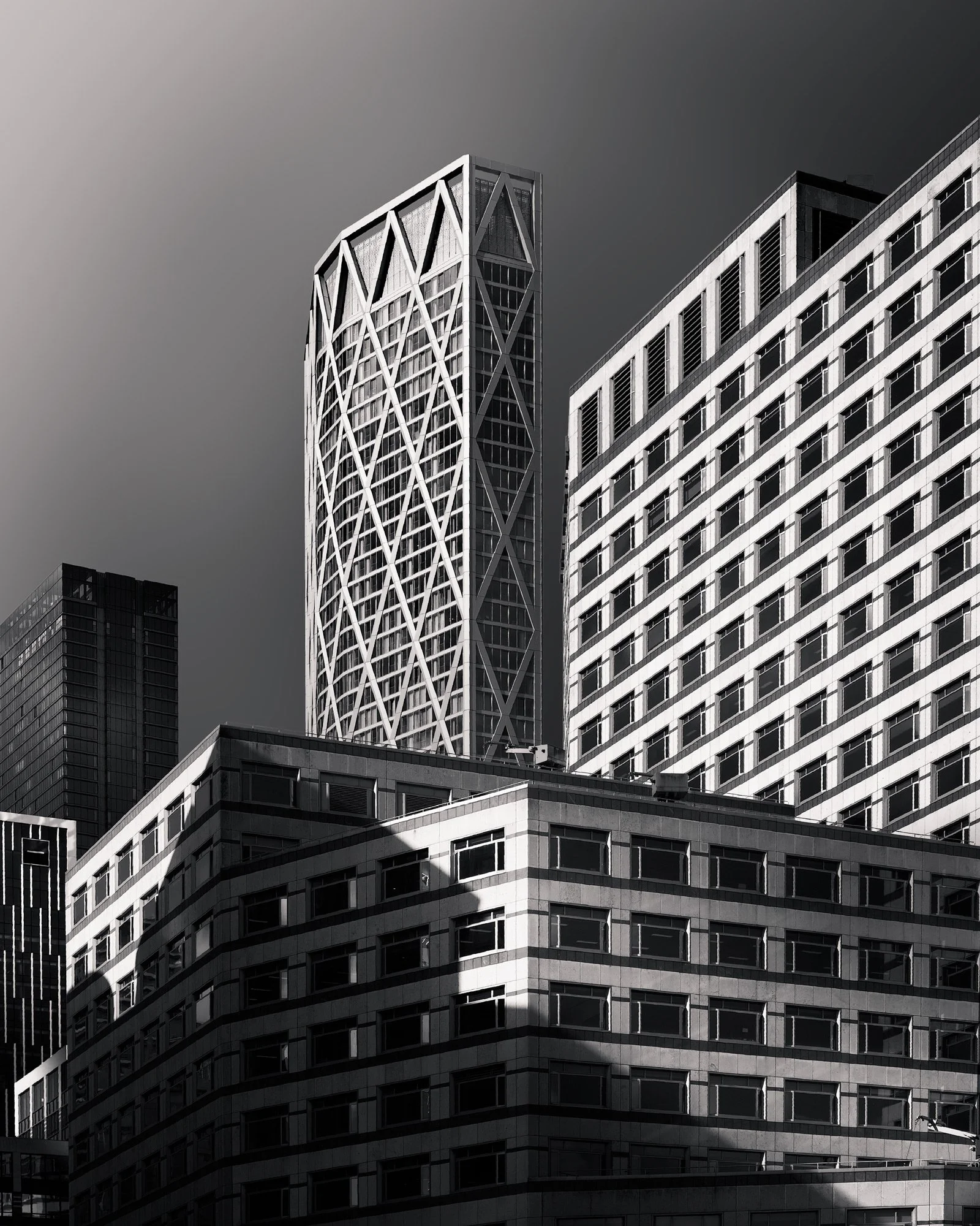 [architecture] [uk] [london] [blackandwhite] Newfoundland Tower Canary Wharf London, diagonal lattice exoskeleton rising above surrounding lower office blocks, sharp tonal contrast. Architectural Photography by Jim Scott.