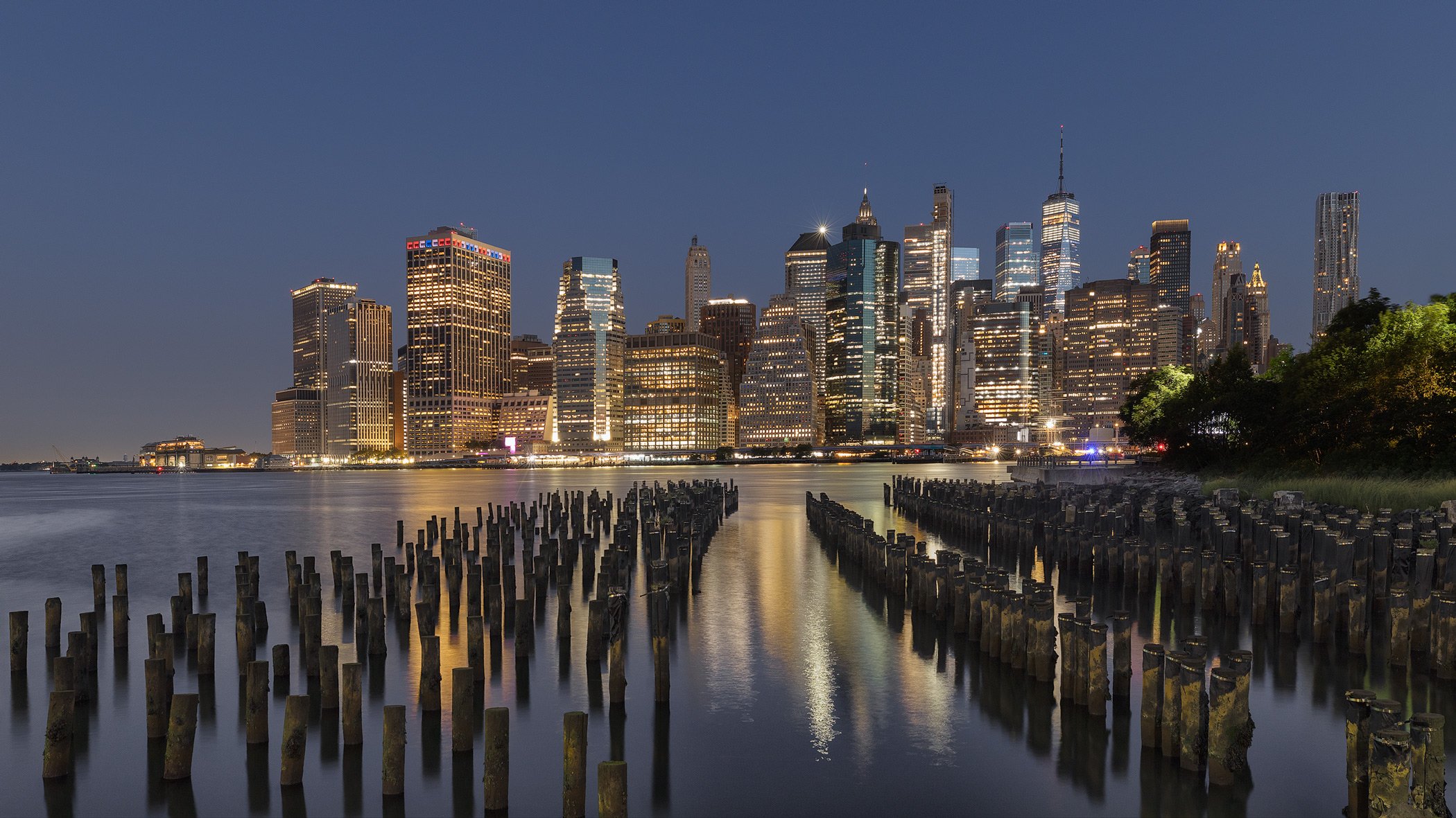 Contemporary architecture in New York City: The Lower Manhattan skyline and One World Trade Center viewed from Brooklyn Bridge Park. A long-exposure night photograph featuring the illuminated skyscrapers of the Financial District reflected in the Eas