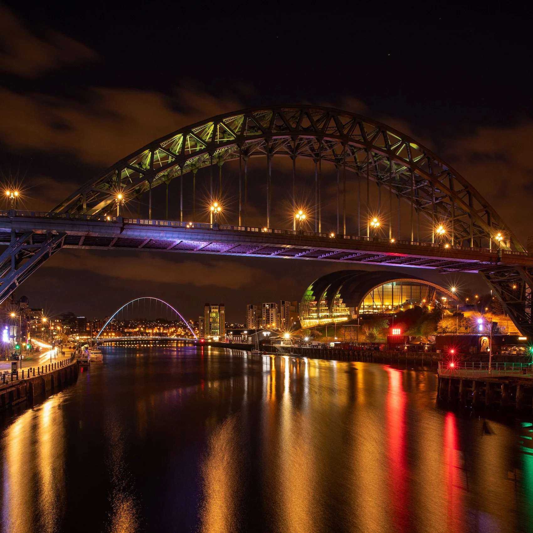 [architecture] [uk] [newcastle] [colour] [square] [nightscape] Tyne Bridge Newcastle at night, green-lit steel arch reflected in the river with the Sage Gateshead glowing beyond. Architectural Photography by Jim Scott.
