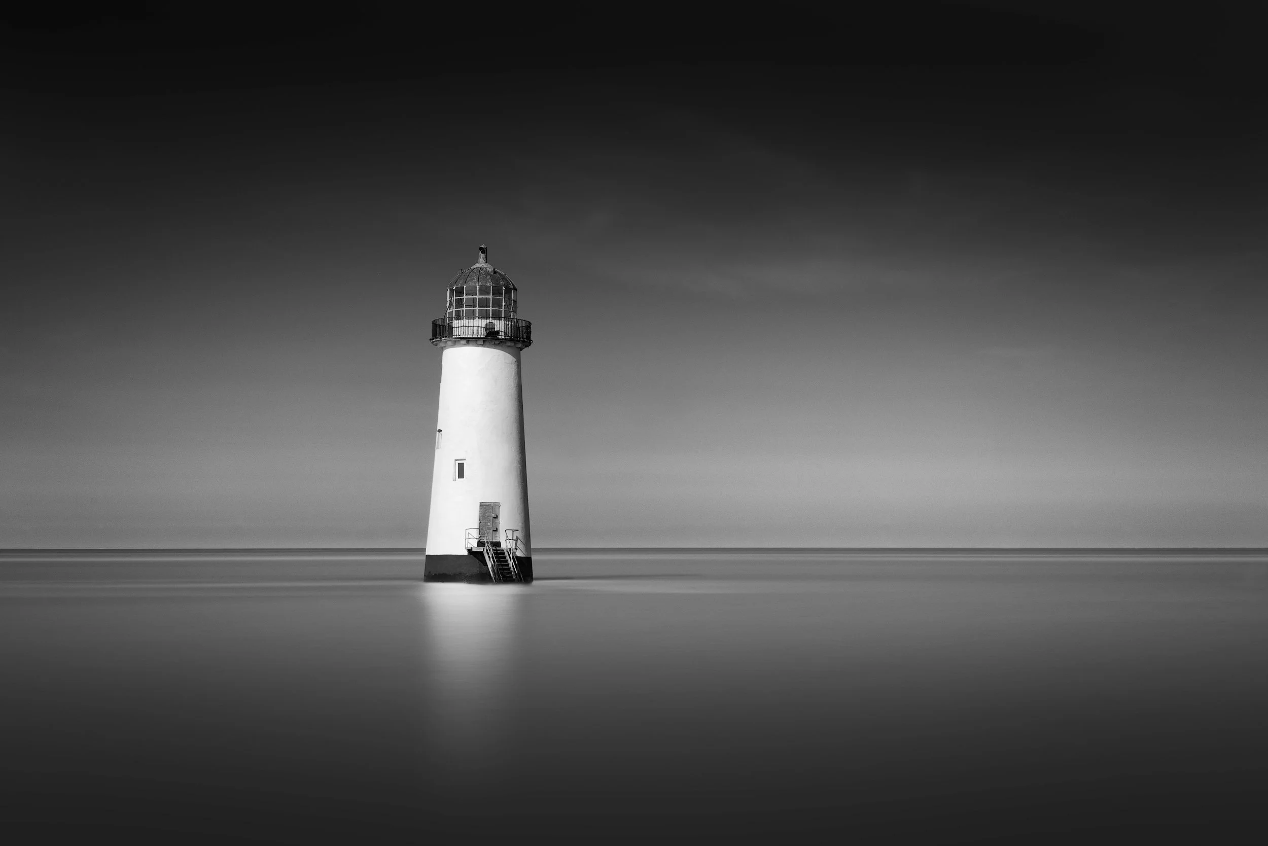 Historic maritime architecture in Talacre, Wales: A minimalist monochromatic study of the Point of Ayr Lighthouse. This architectural photograph captures the abandoned tower rising directly from the Irish Sea. The long-exposure technique creates a pe