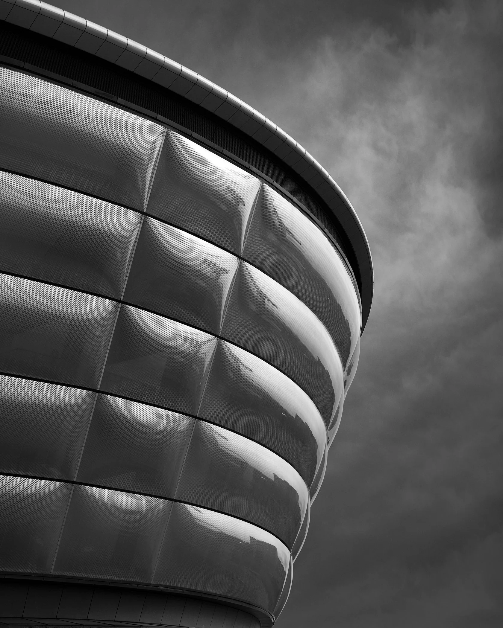 The OVO Hydro arena close-up. A dramatic, low-angle black and white architectural detail capturing the unique EFTE-cushioned facade and mesh panels against a turbulent, stormy sky over the River Clyde.