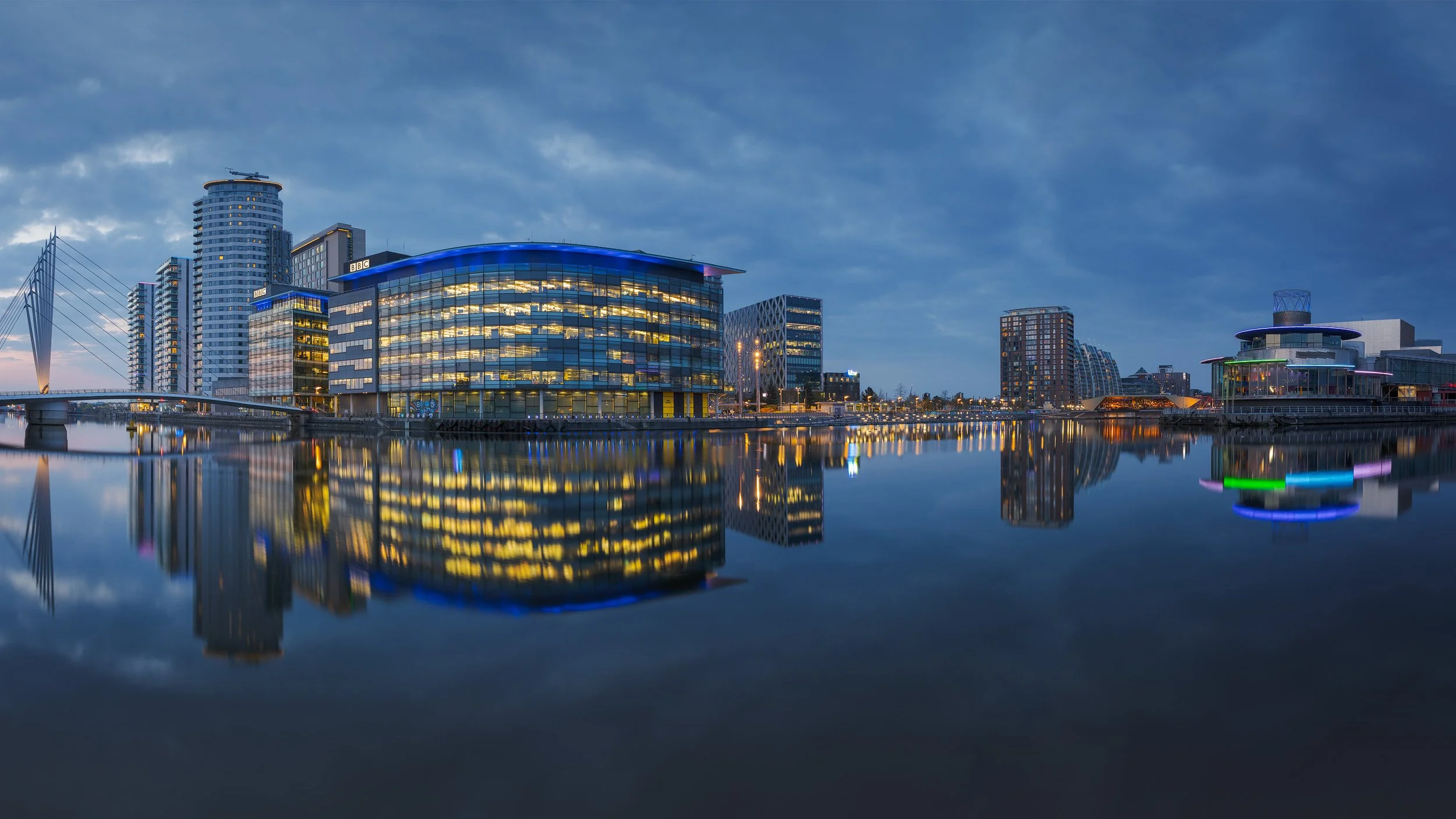 [architecture] [uk] [salford] [colour] [panoramic] [nightscape] Blue hour panorama of MediaCityUK Salford, BBC building glowing gold across still water with the Lowry Theatre beyond. Architectural Photography by Jim Scott.