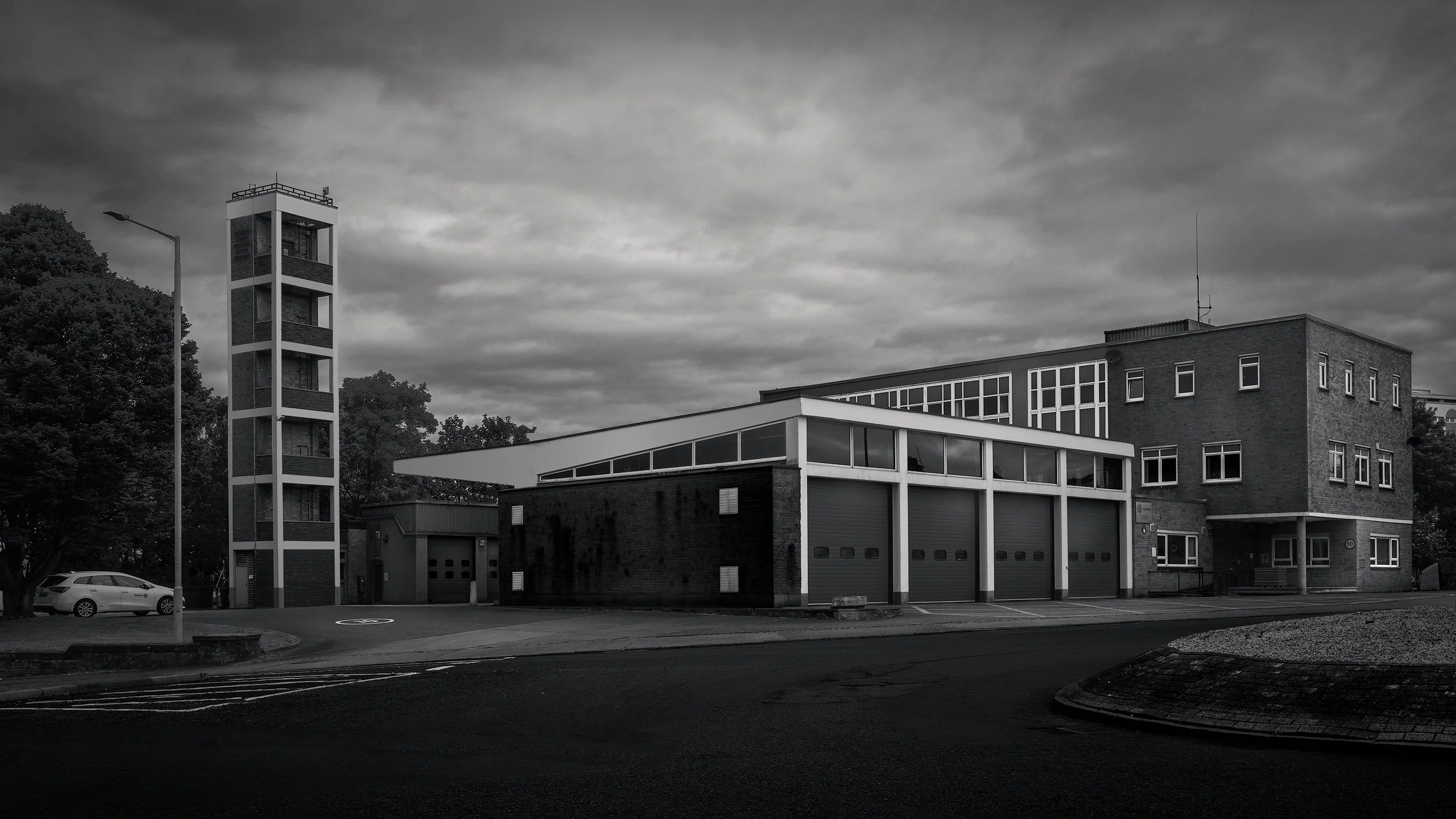 Mid-century civic architecture in Ayr, Scotland: A dramatic monochromatic study of the Ayr Fire Station. This architectural photograph captures the utilitarian geometry of the station’s main bays and its iconic gridded hose tower. The high-contrast b