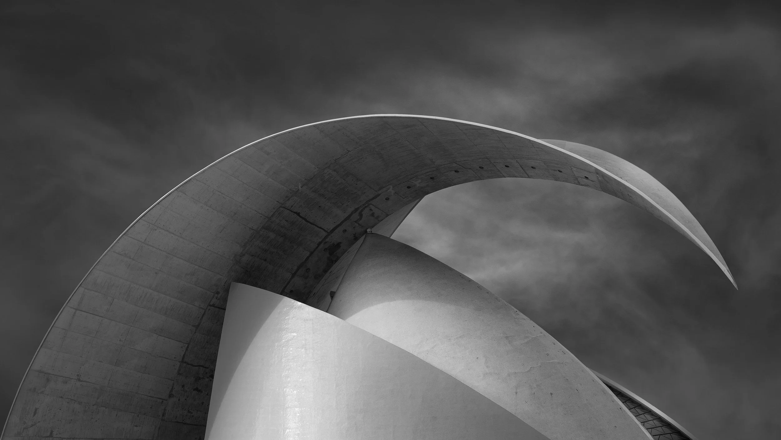 [architecture] [europe] [santacruzdetenerife] [blackandwhite] [panoramic] Auditorio de Tenerife roof in landscape, the concrete crescent arch and white shell interplay under a moody grey sky. Architectural Photography by Jim Scott.