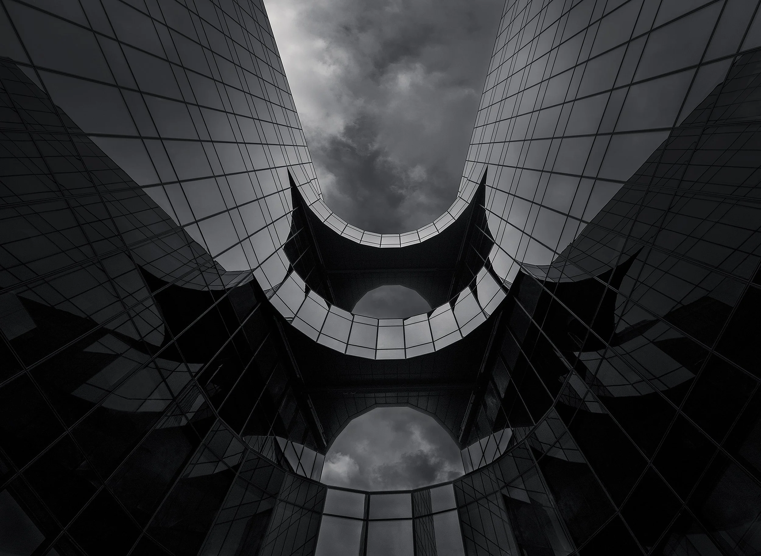 [architecture] [uk] [london] [blackandwhite] PwC More London, looking up through curved glass arch between twin towers forming batman silhouette, dark dramatic black and white. Architectural Photography by Jim Scott.